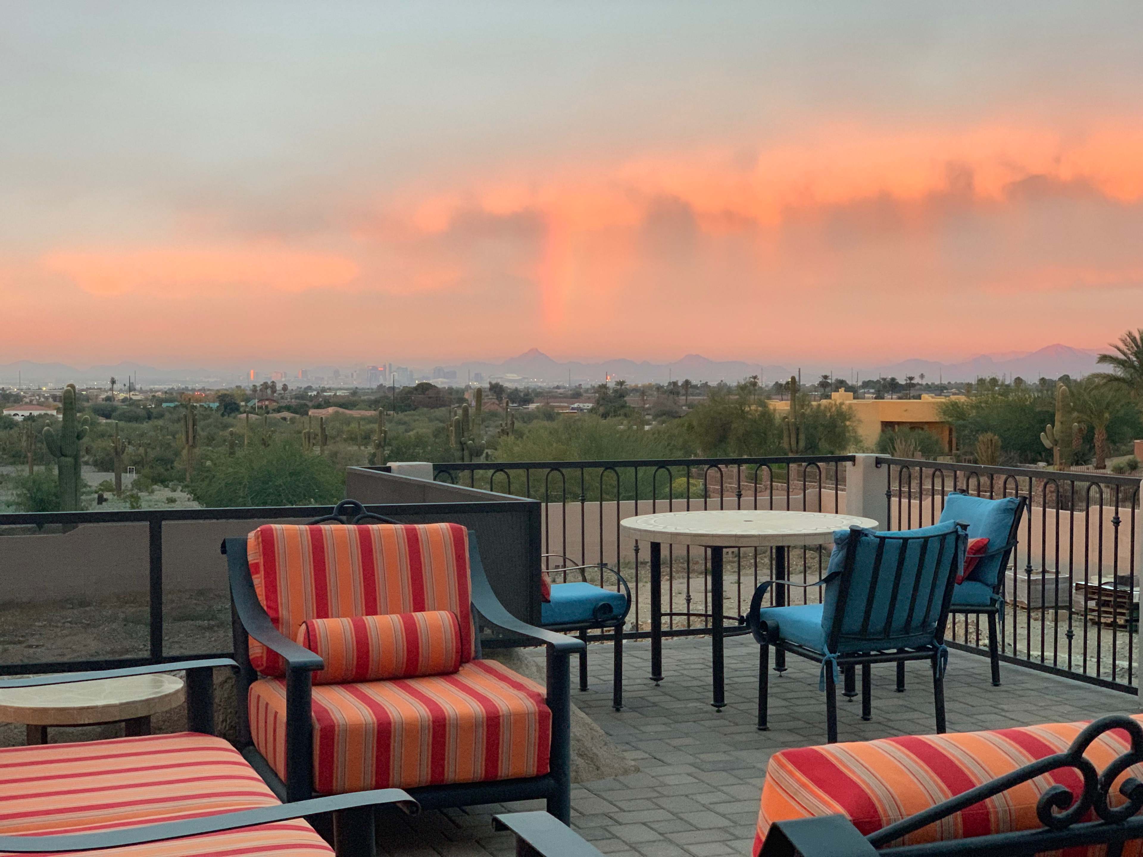 The image shows a patio with striped chairs and a table overlooking a sunset-lit landscape featuring distant mountains and a city skyline.