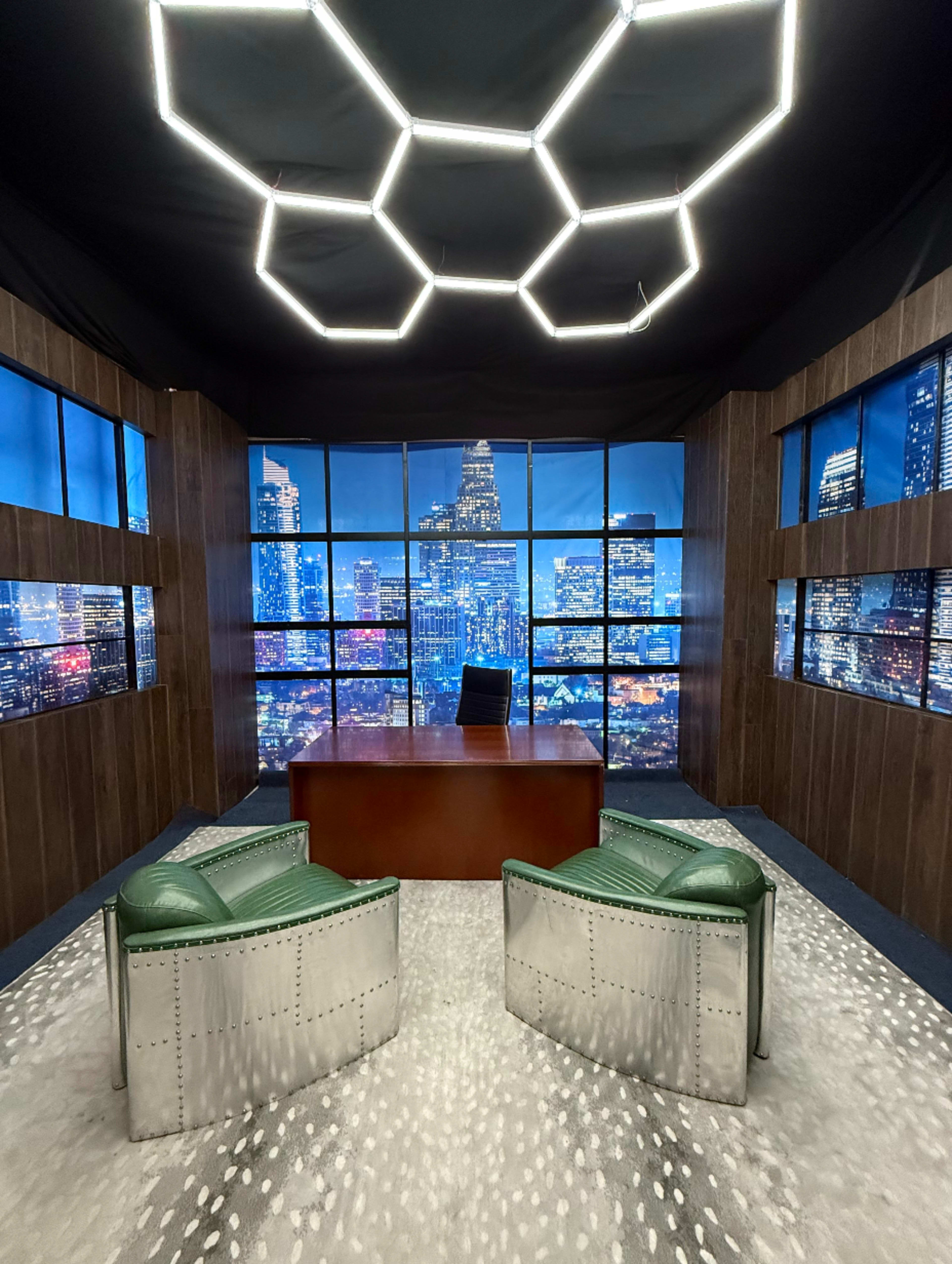 A modern office space features two green chairs facing a desk, with large windows showcasing a city skyline at night.