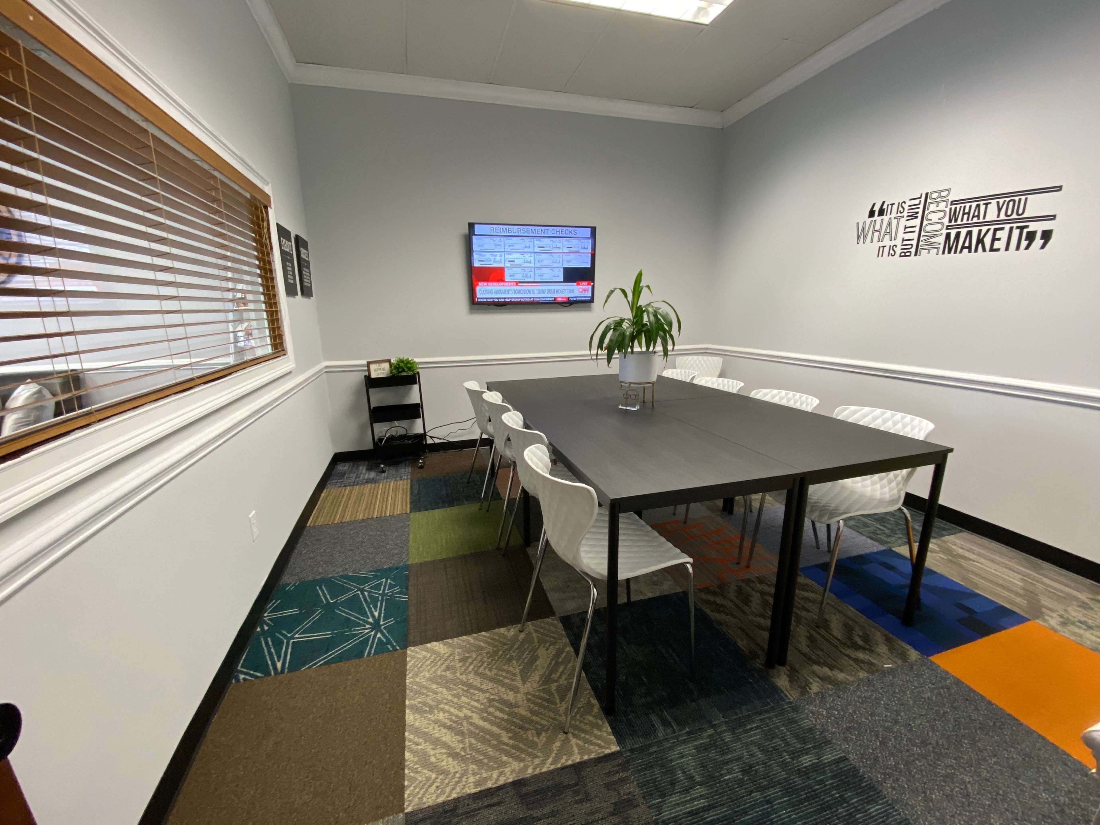 The image shows a modern conference room with a rectangular table surrounded by white chairs, a television on the wall, and a decorative plant.