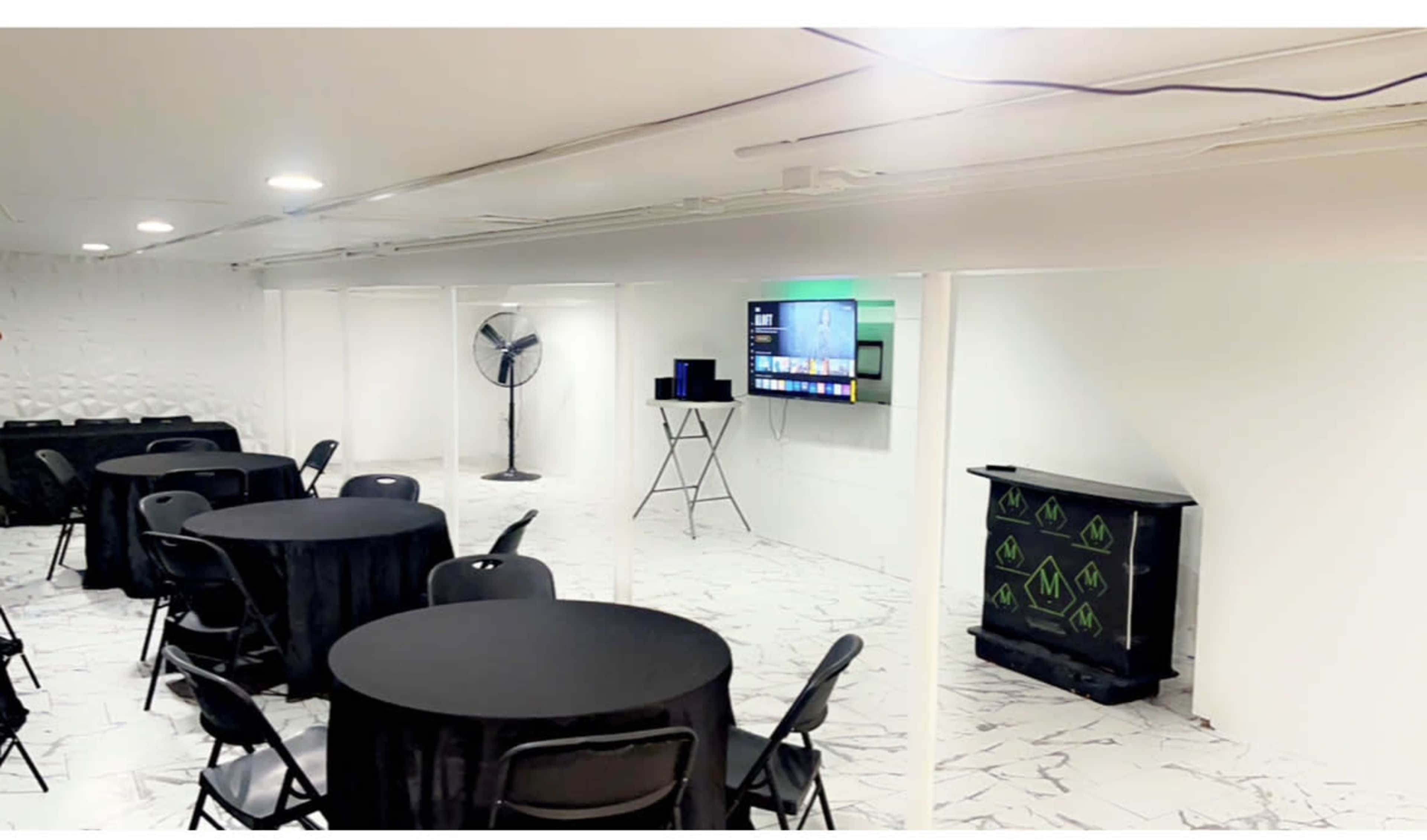 A clean, white-walled meeting space with several black tables and chairs, a fan, and a television mounted on the wall.