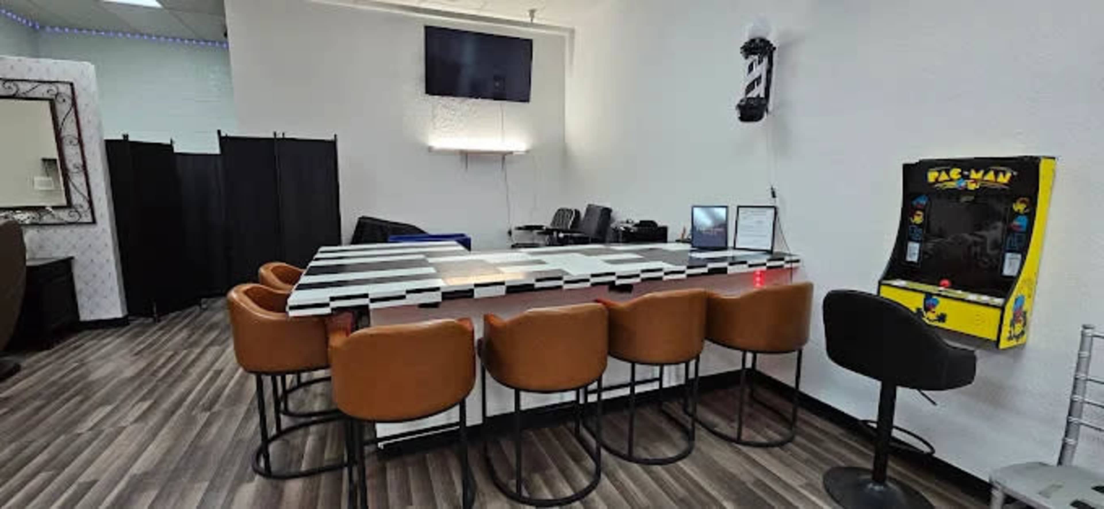The image shows a modern barbershop interior featuring a striped countertop with several brown barstools, a vintage arcade game, and a television on the wall.