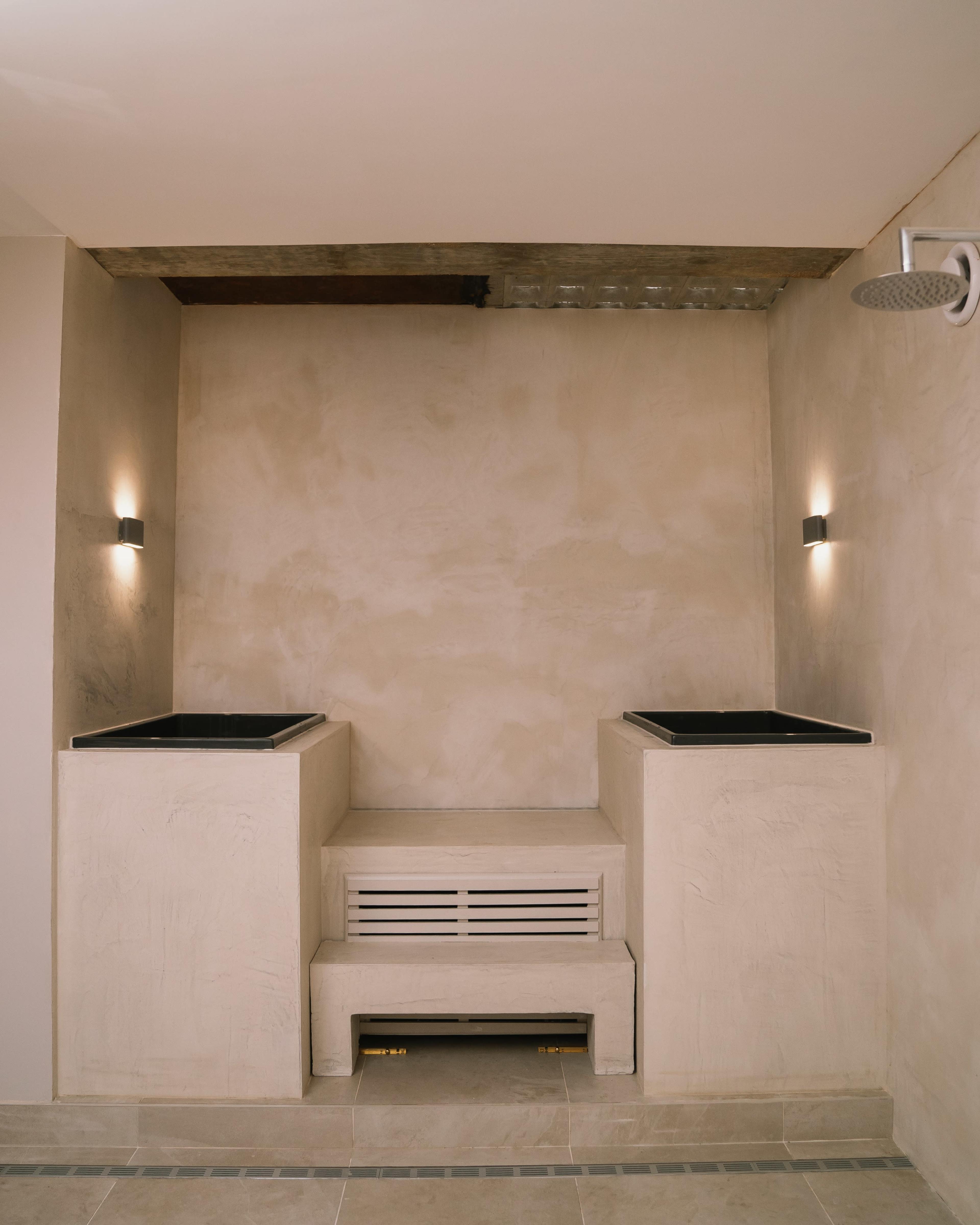 A minimalistic bathroom area featuring two square black sinks, a central vent, and wall-mounted lighting in a textured beige wall setting.