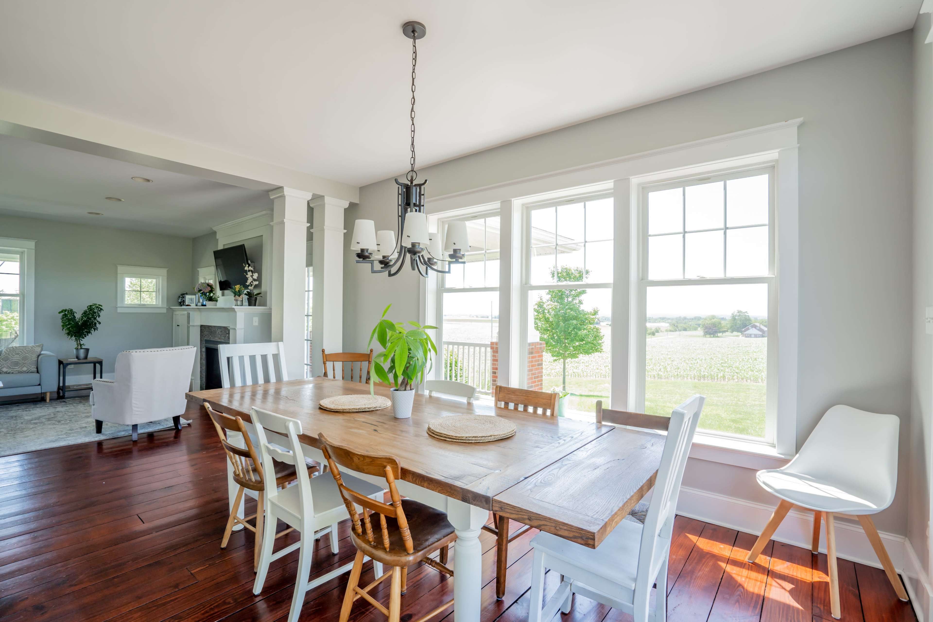 A wooden dining table is set with placemats and surrounded by various chairs, with large windows overlooking a field.
