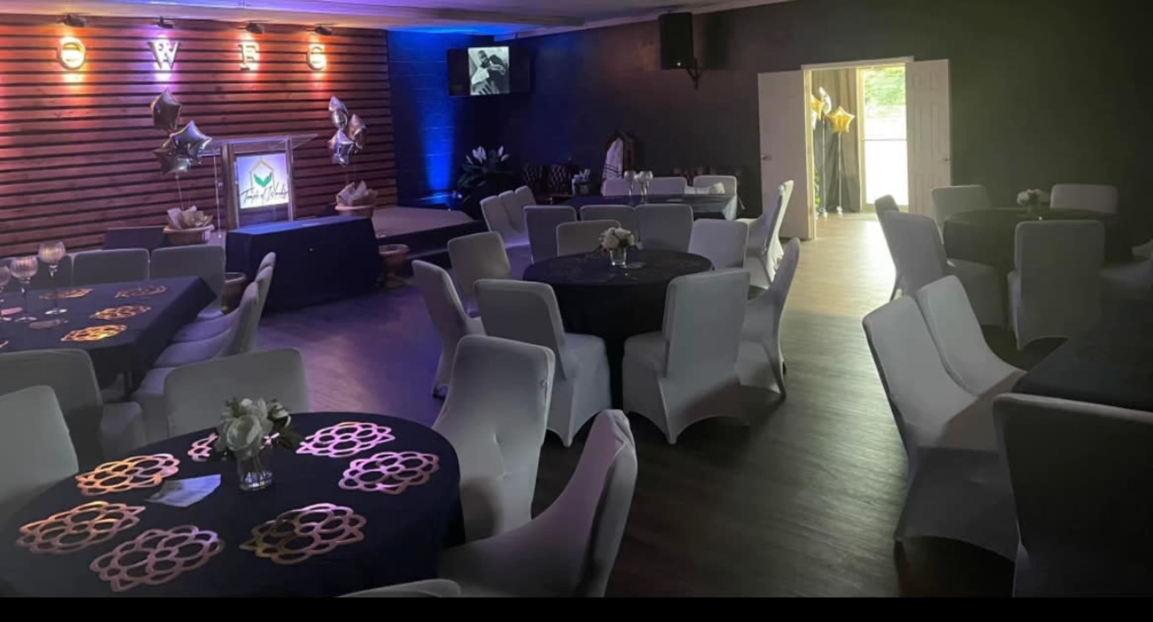A spacious event room set up with round tables adorned with dark tablecloths and decorative centerpieces, alongside elegant white chairs, illuminated by colorful lighting.
