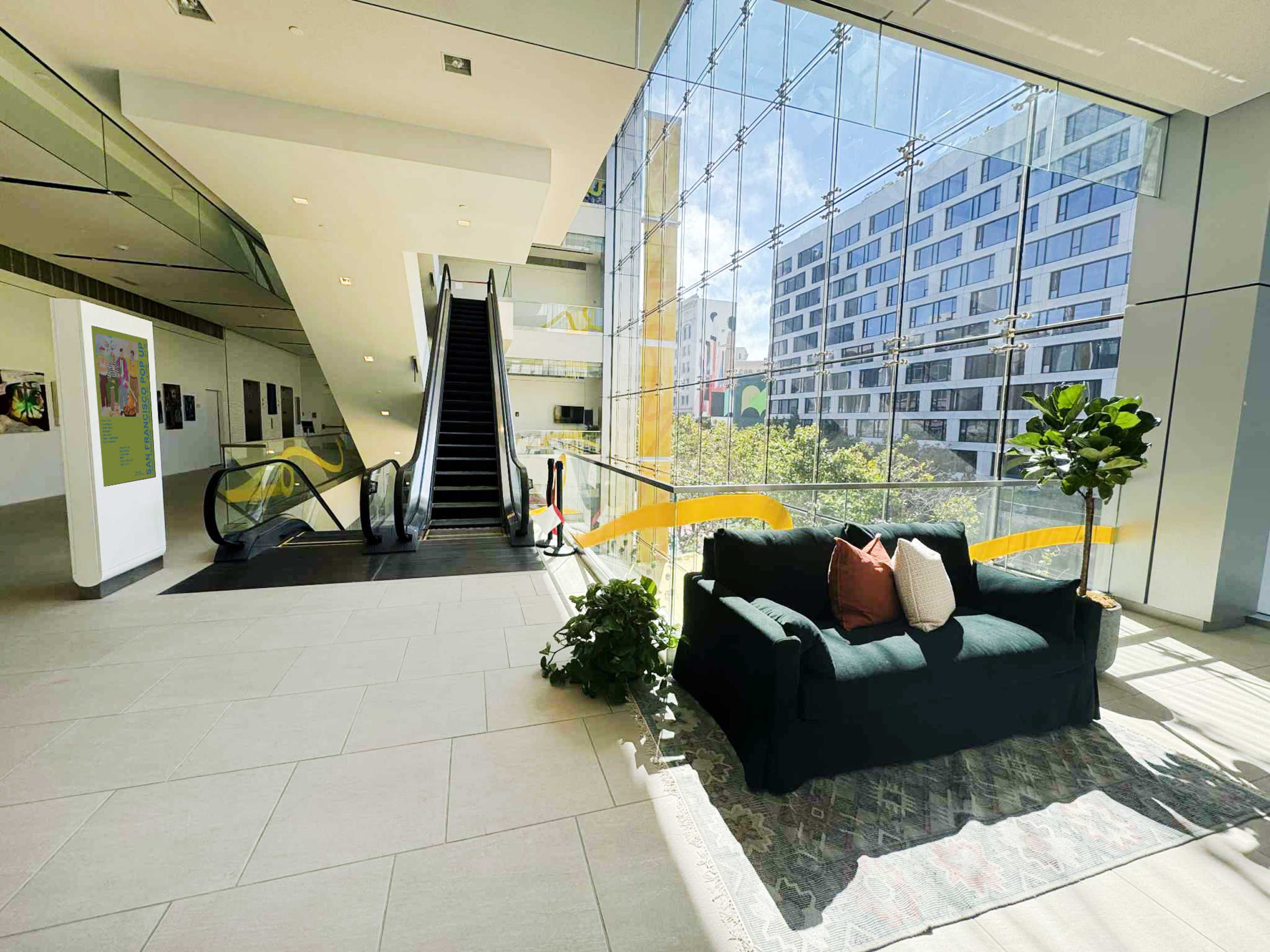 The image shows a modern lobby with a seating area featuring a dark couch and decorative pillows, large windows allowing natural light, and an escalator leading to a higher floor.