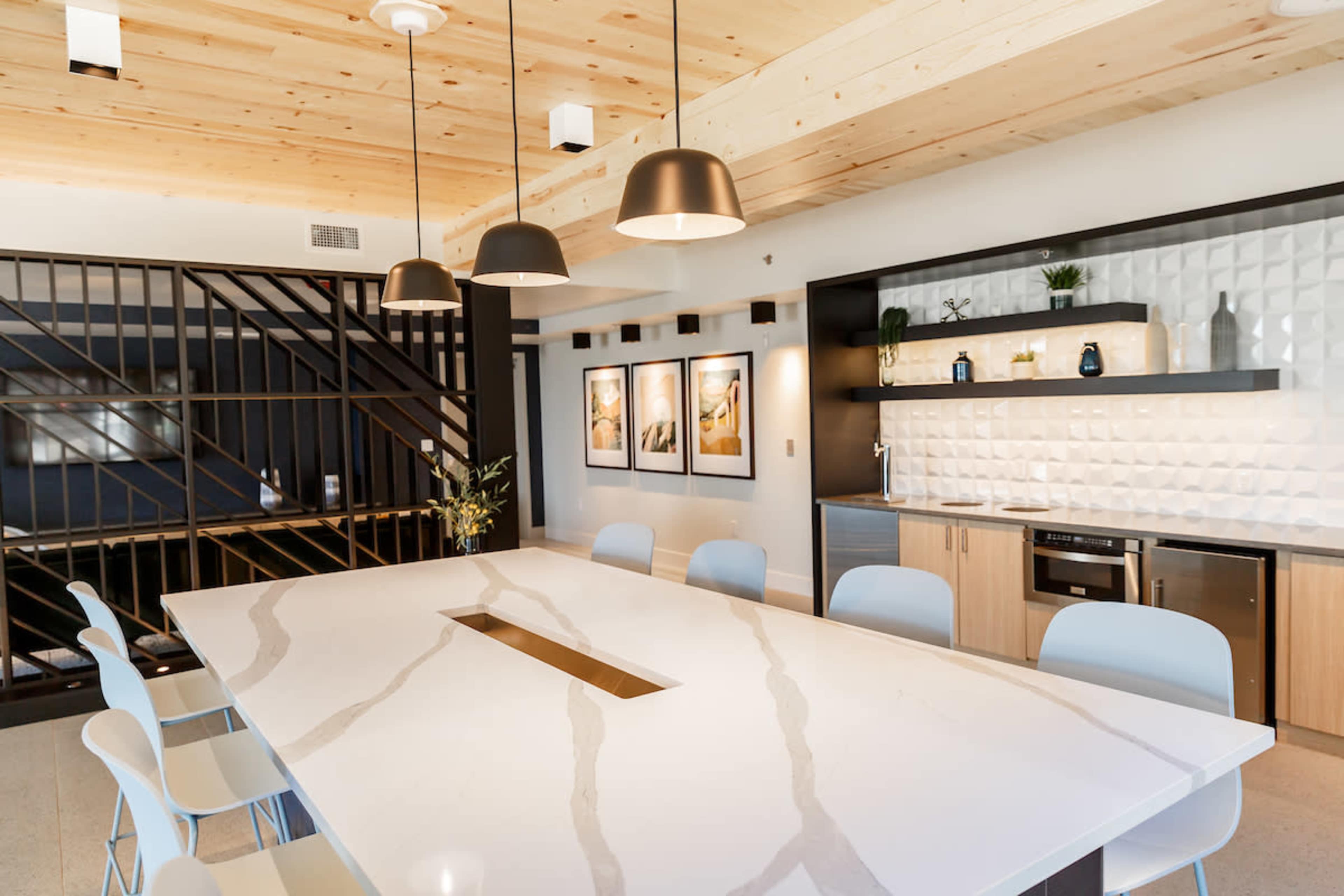 A modern kitchen with a large central table, minimalist cabinetry, and a wall adorned with framed artwork, illuminated by hanging pendant lights.