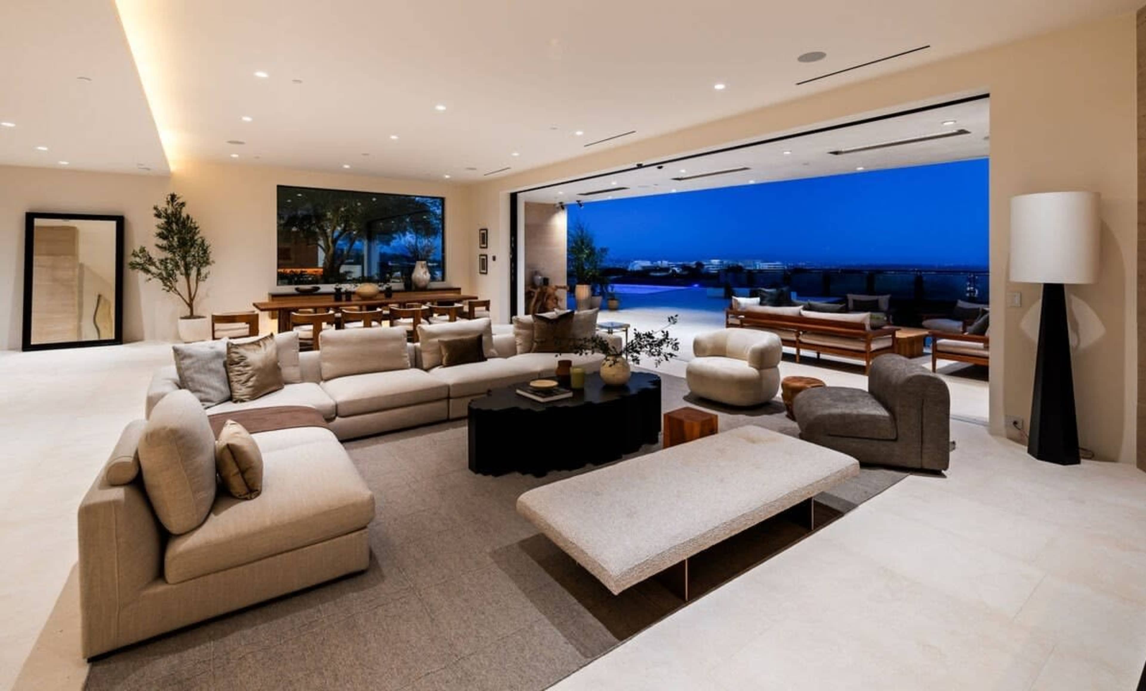 The image shows a modern, open-concept living room with a large sectional sofa, a circular coffee table, and panoramic views of an outdoor area lit by dusk.