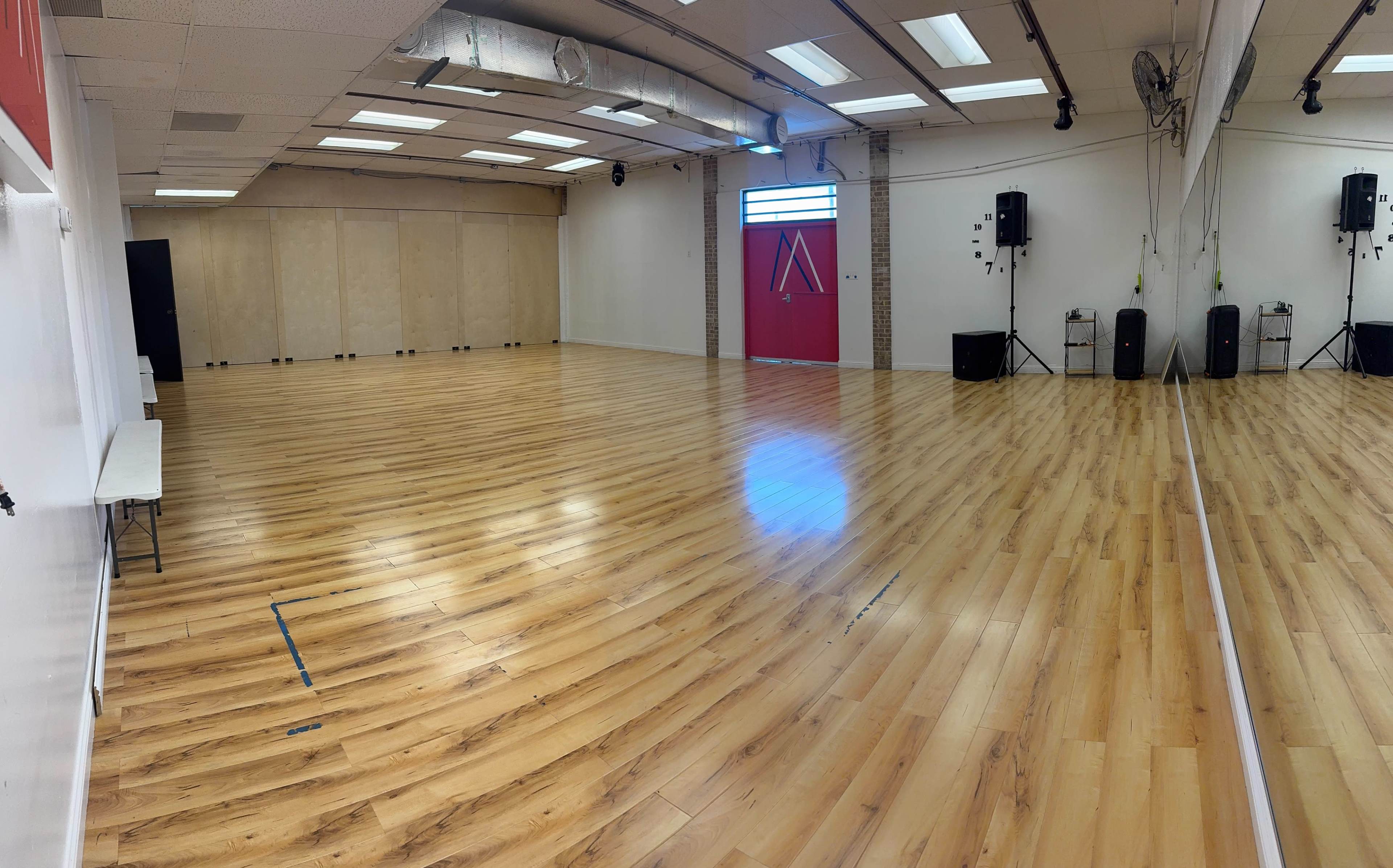 The image shows an empty dance studio with wooden flooring, mirrored walls, and a red door at one end.