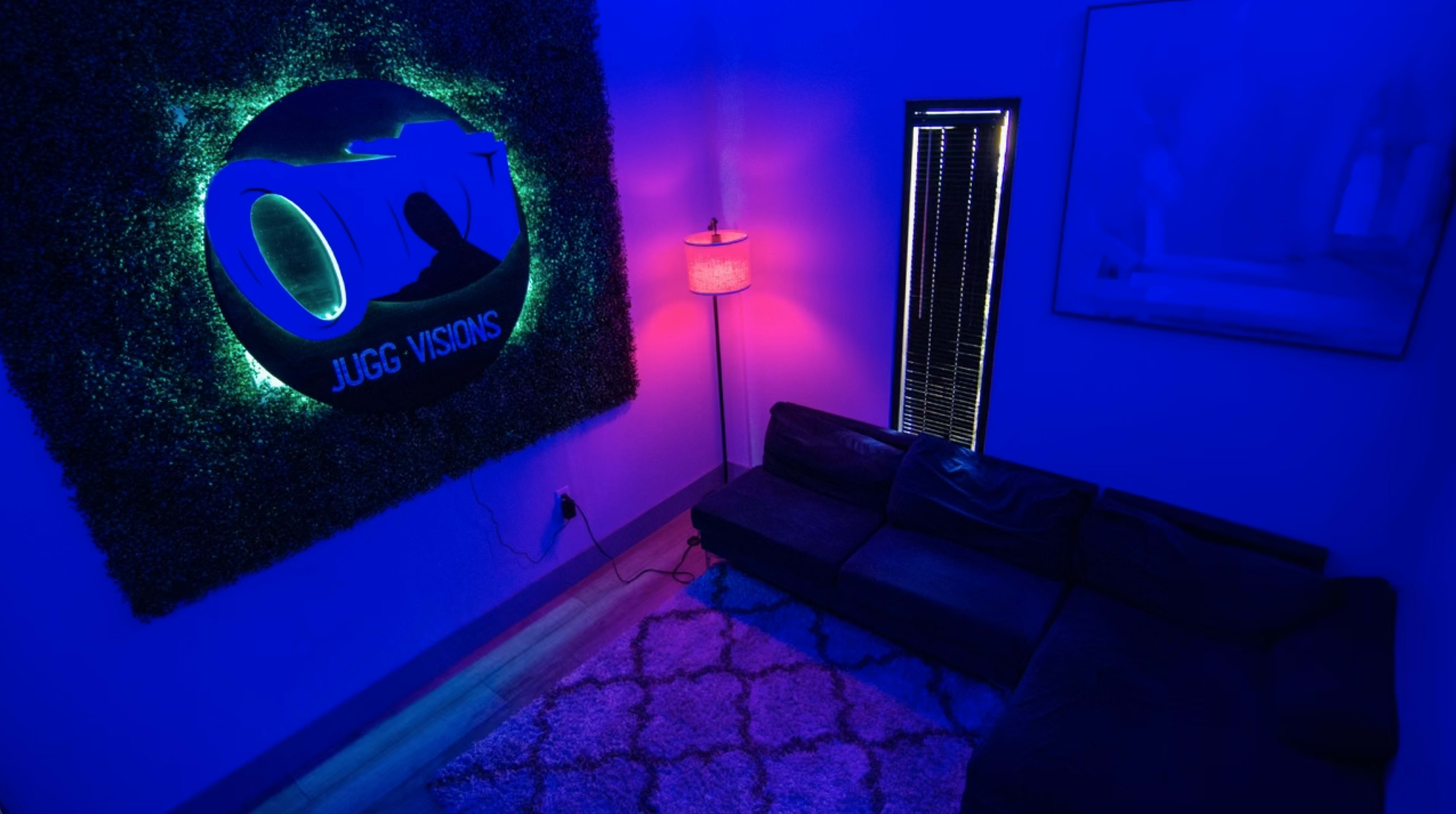 A modern room with a dark couch, a patterned rug, and a neon sign on the wall surrounded by greenery, illuminated by colorful lights.