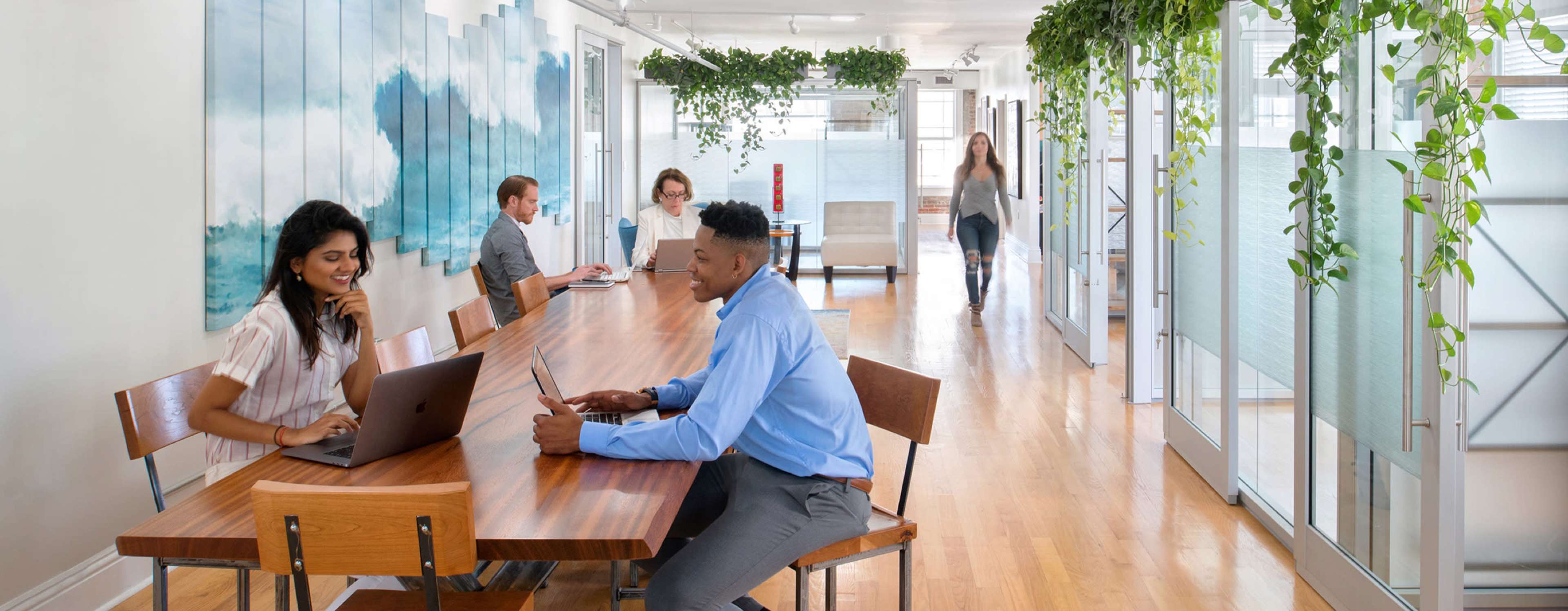 A diverse group of people works at a long wooden table in a modern office space with greenery and natural light.