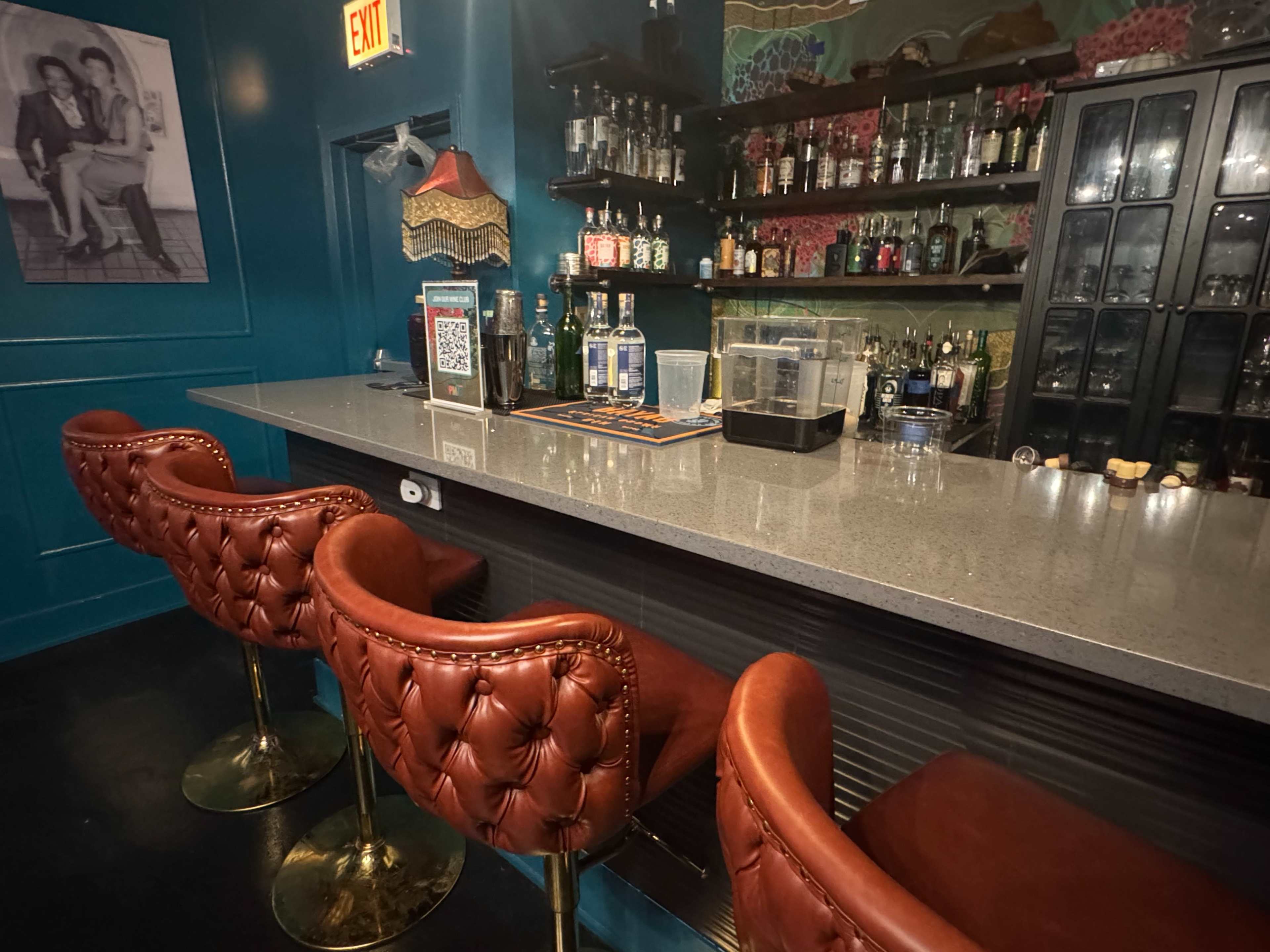 The image shows a stylish bar area with a polished countertop, three red leather barstools, and a variety of bottles displayed on shelves in a dimly lit environment.