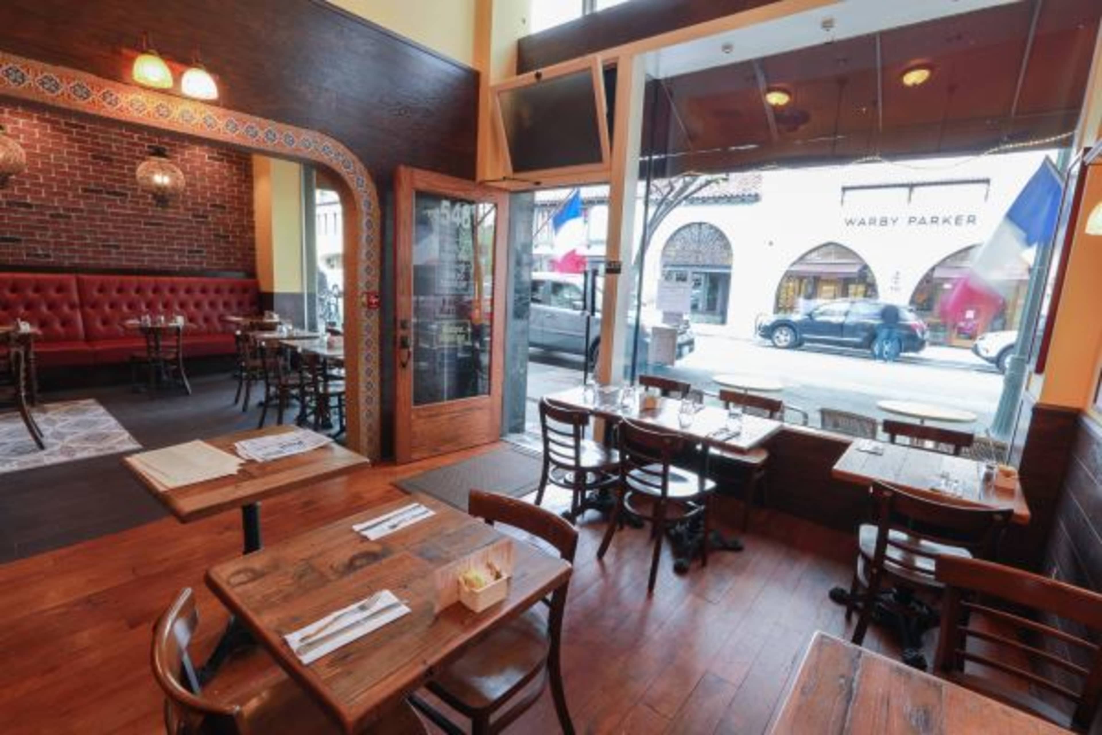 The interior of a restaurant featuring wooden tables and chairs, red upholstered seating, and large windows overlooking a street with visible storefronts.