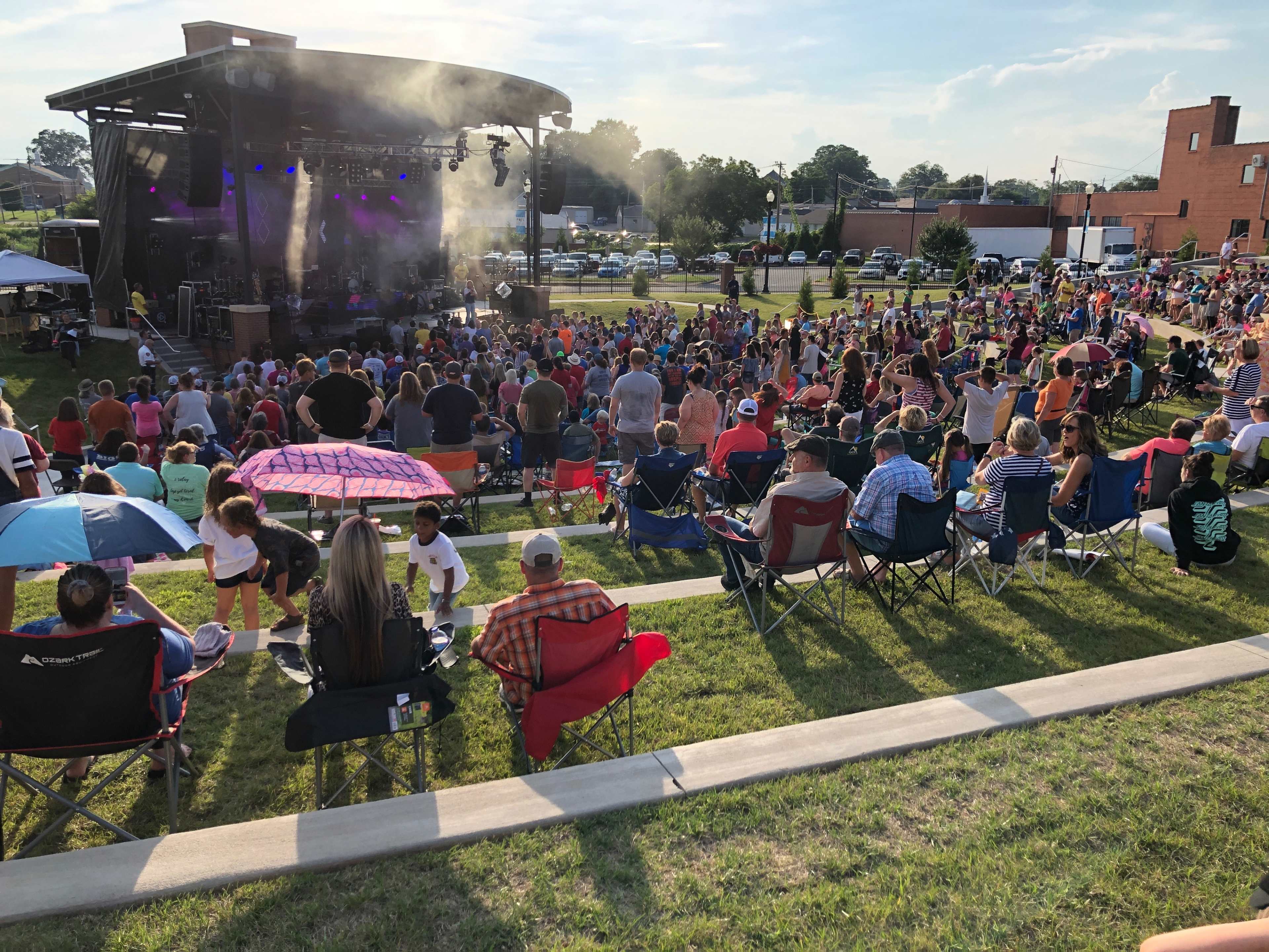 A large crowd gathers on a grassy area in front of a stage with performers, surrounded by food vendors and tents.