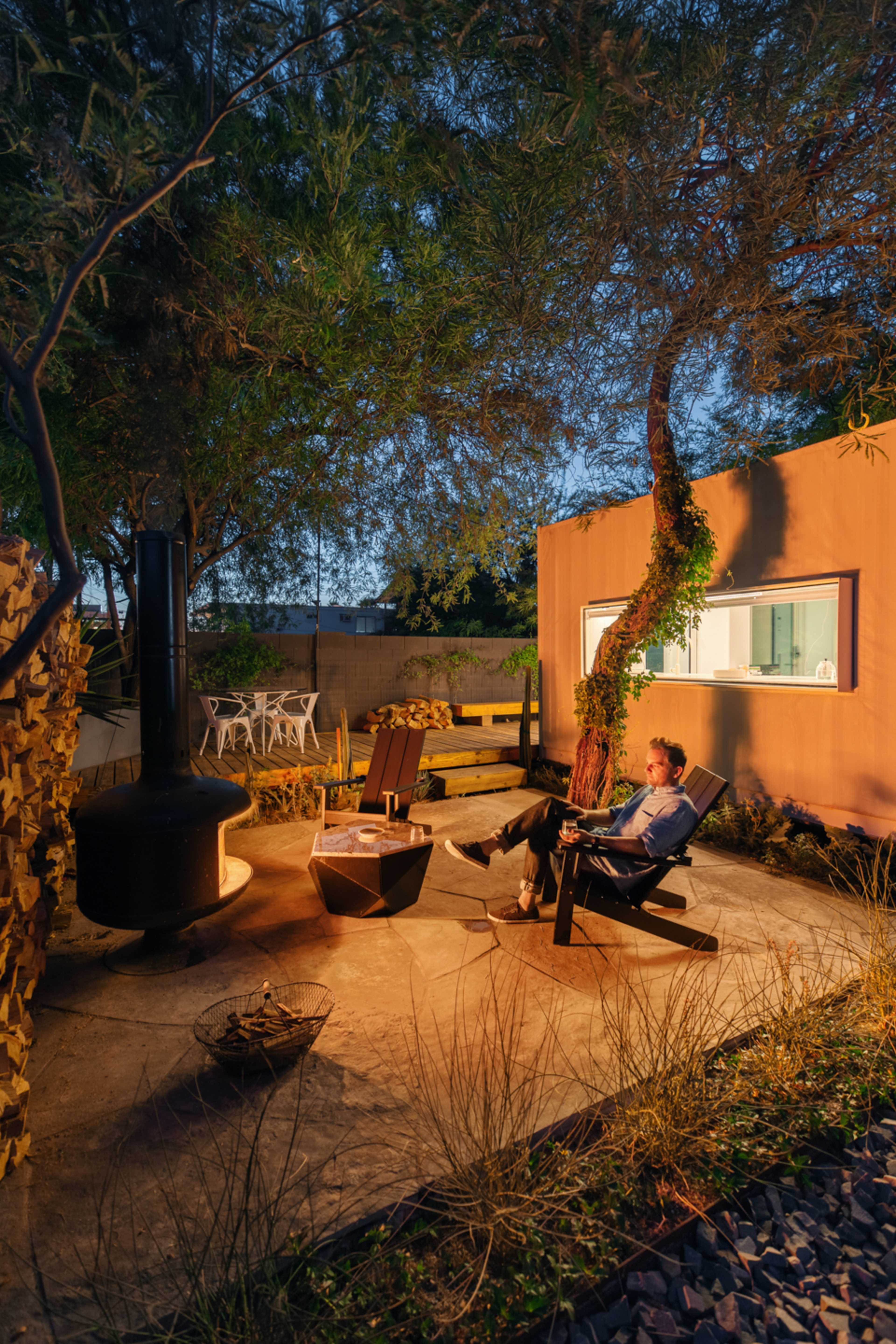A man sits in an outdoor seating area beside a fire pit, surrounded by trees and a dimly-lit wall, with a small table and chairs visible in the background.