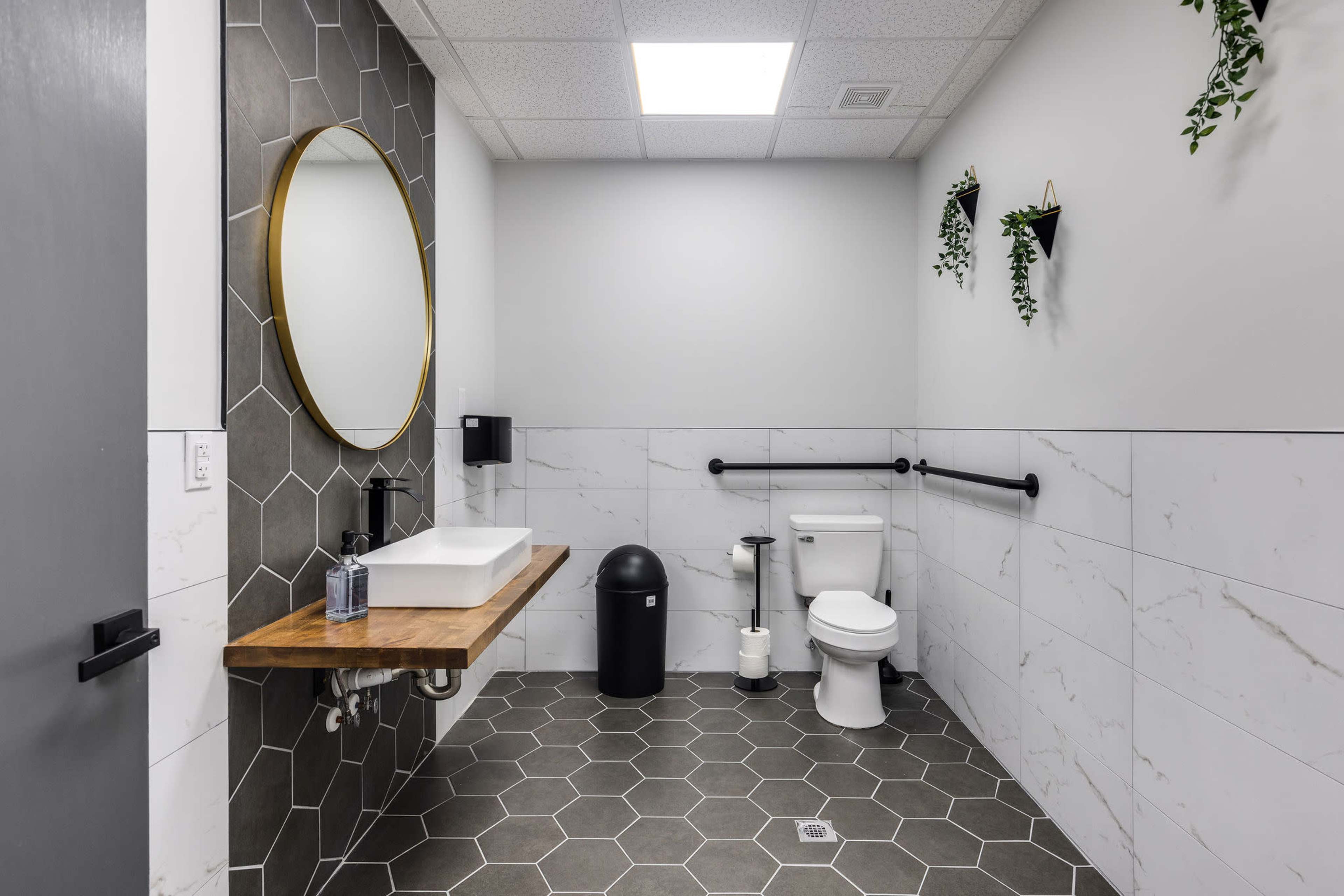 A modern bathroom featuring a circular mirror, a wooden countertop with a sink, a toilet, and hexagonal tile flooring, along with wall-mounted plants.