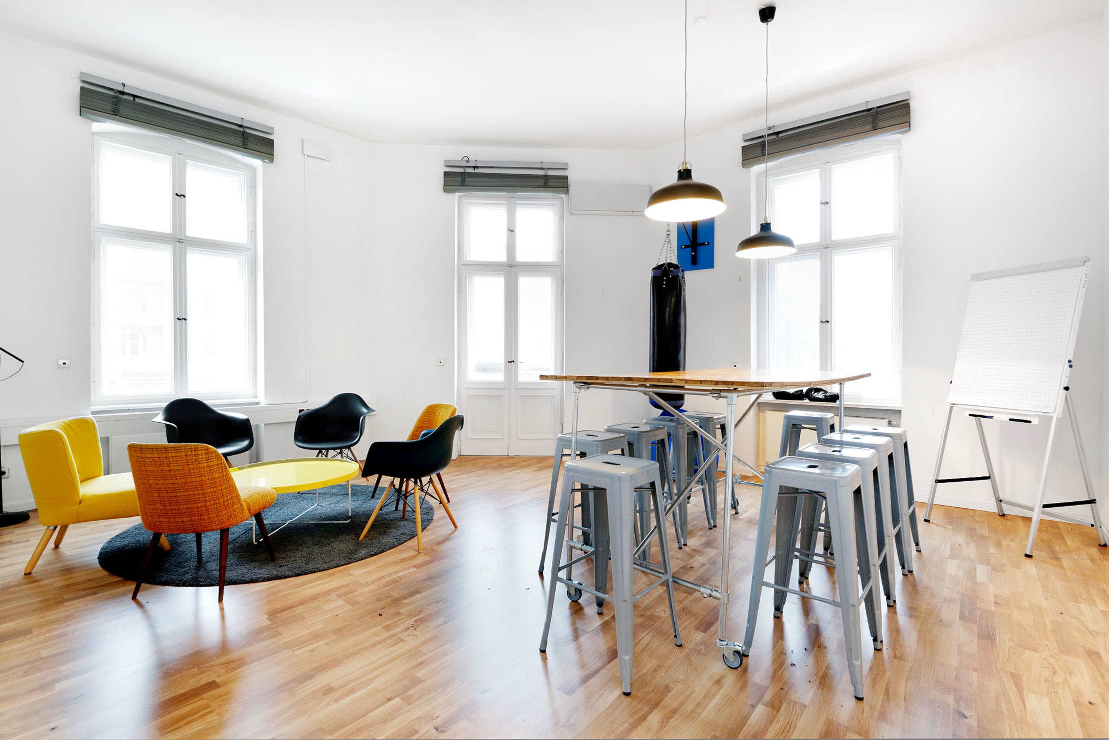 A modern meeting room with a large round carpet, a high table surrounded by stools, colorful seating options, a flip chart, and large windows letting in natural light.