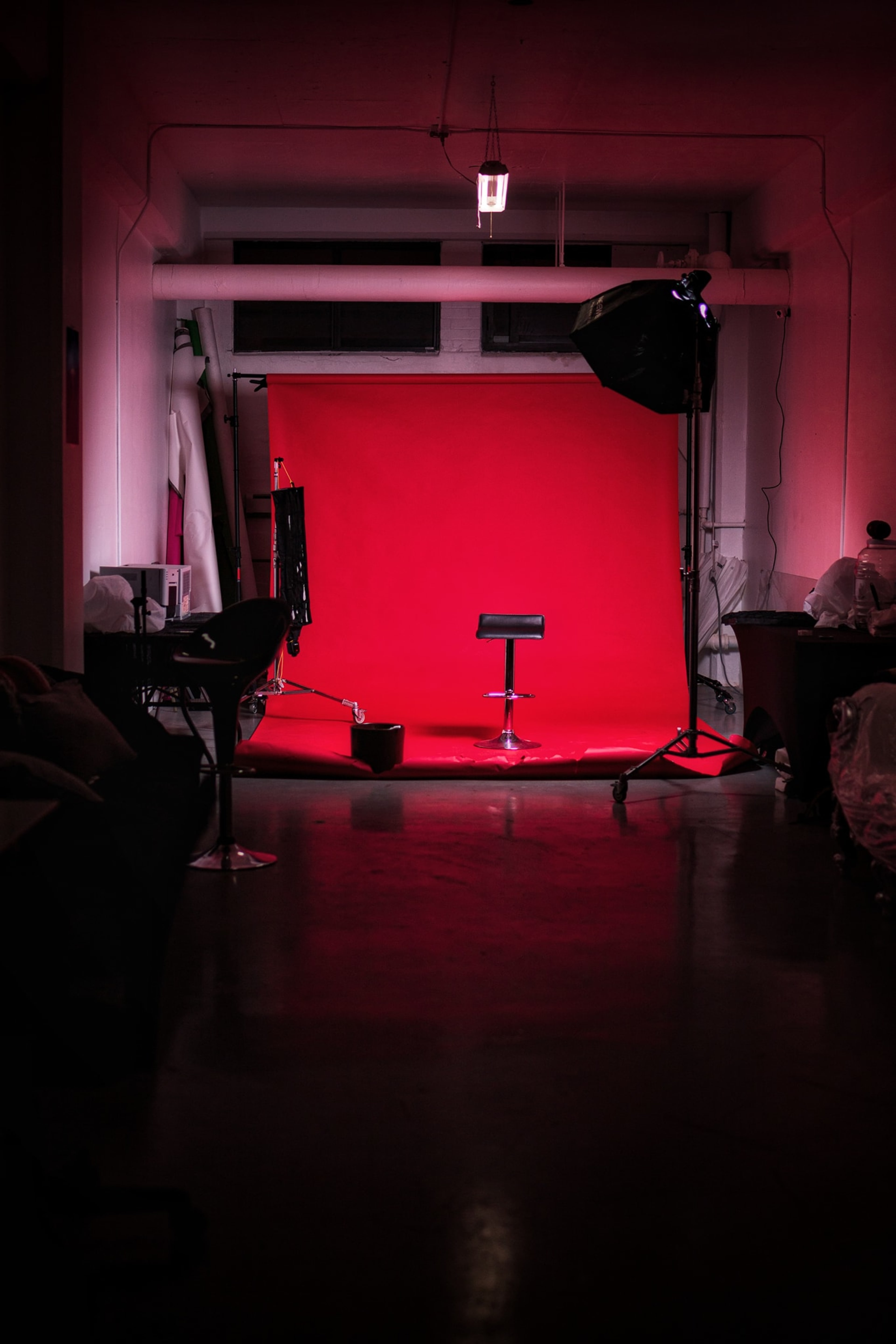 A photography studio features a red backdrop with a black stool and lighting equipment set up for a shoot.