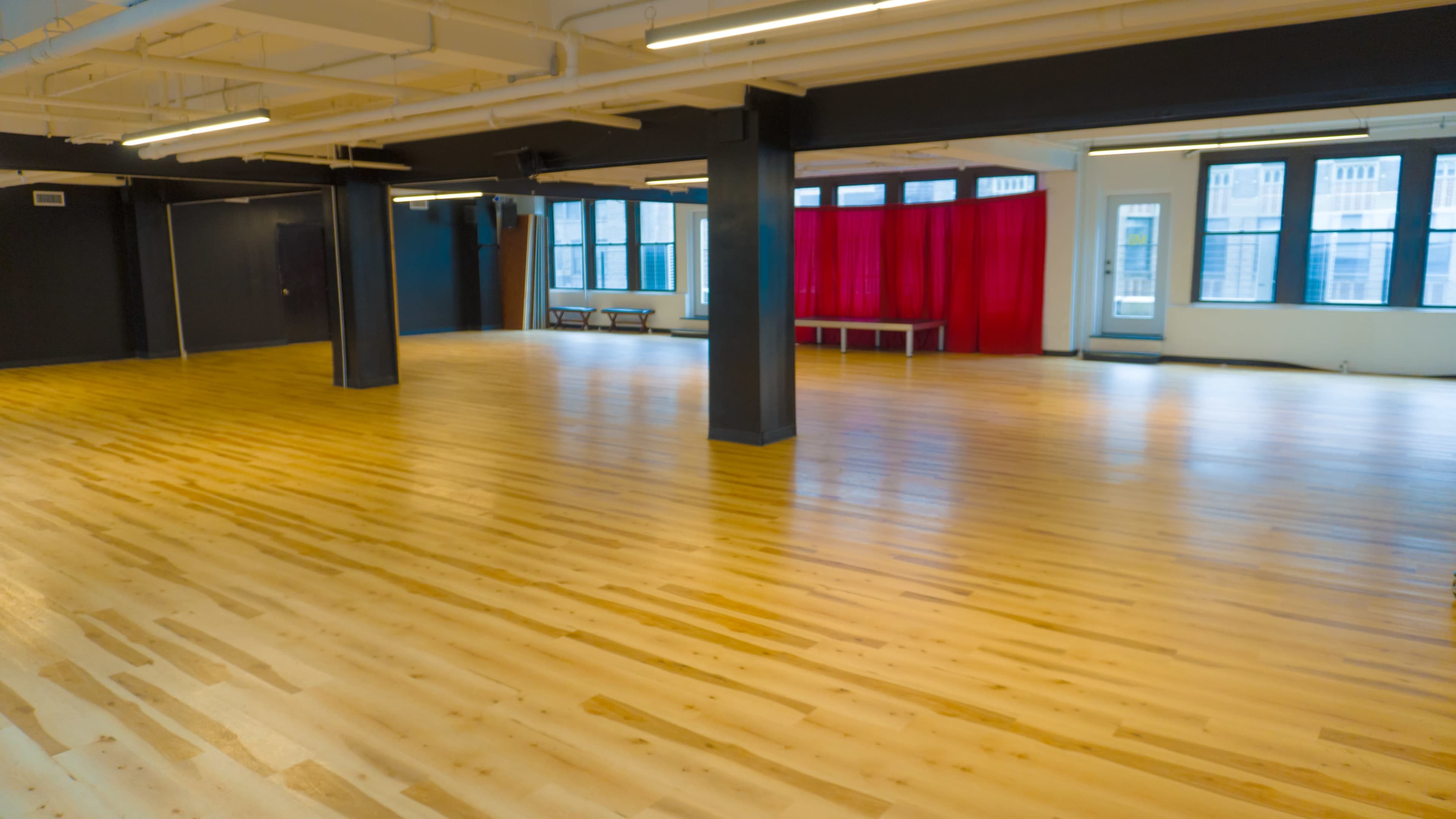 The image shows a spacious room with polished wooden flooring, large windows, and a red curtain on one side.