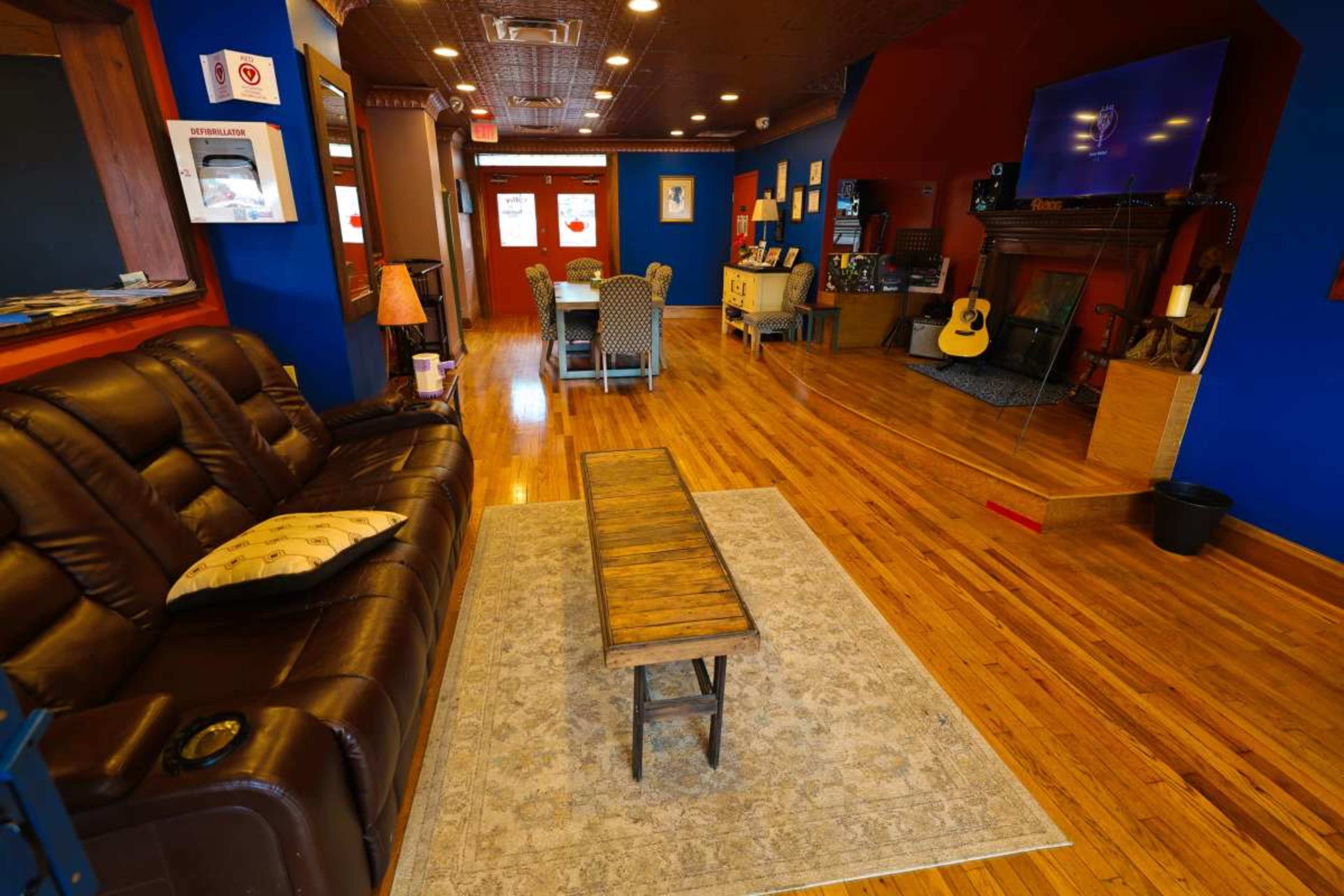 The image shows a cozy room with a brown leather couch, a wooden coffee table, a guitar, and a dining area with a blue and red color scheme.