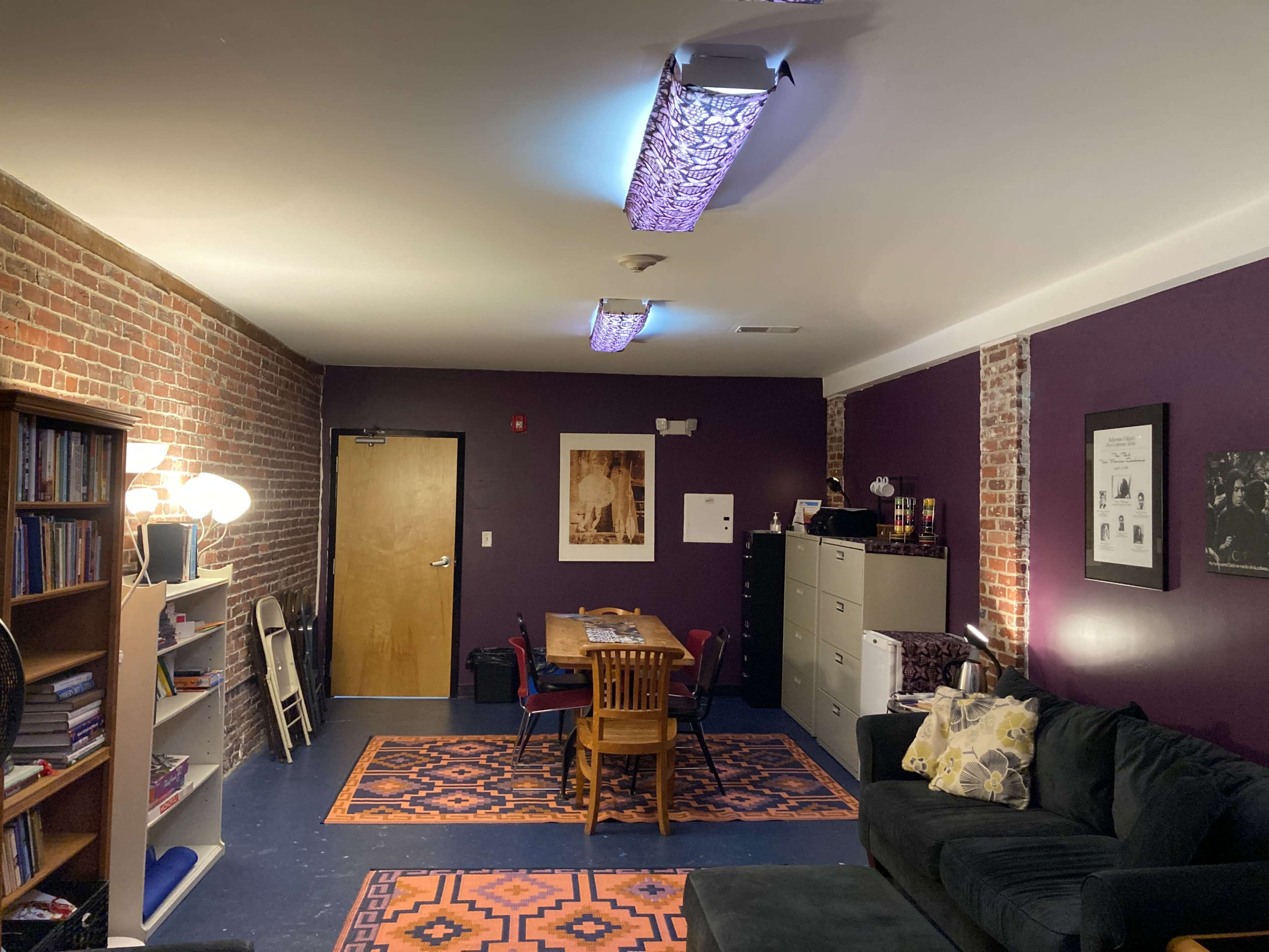 The image shows a room with purple walls, a brick wall section, a table with chairs, a couch, and shelving filled with books.