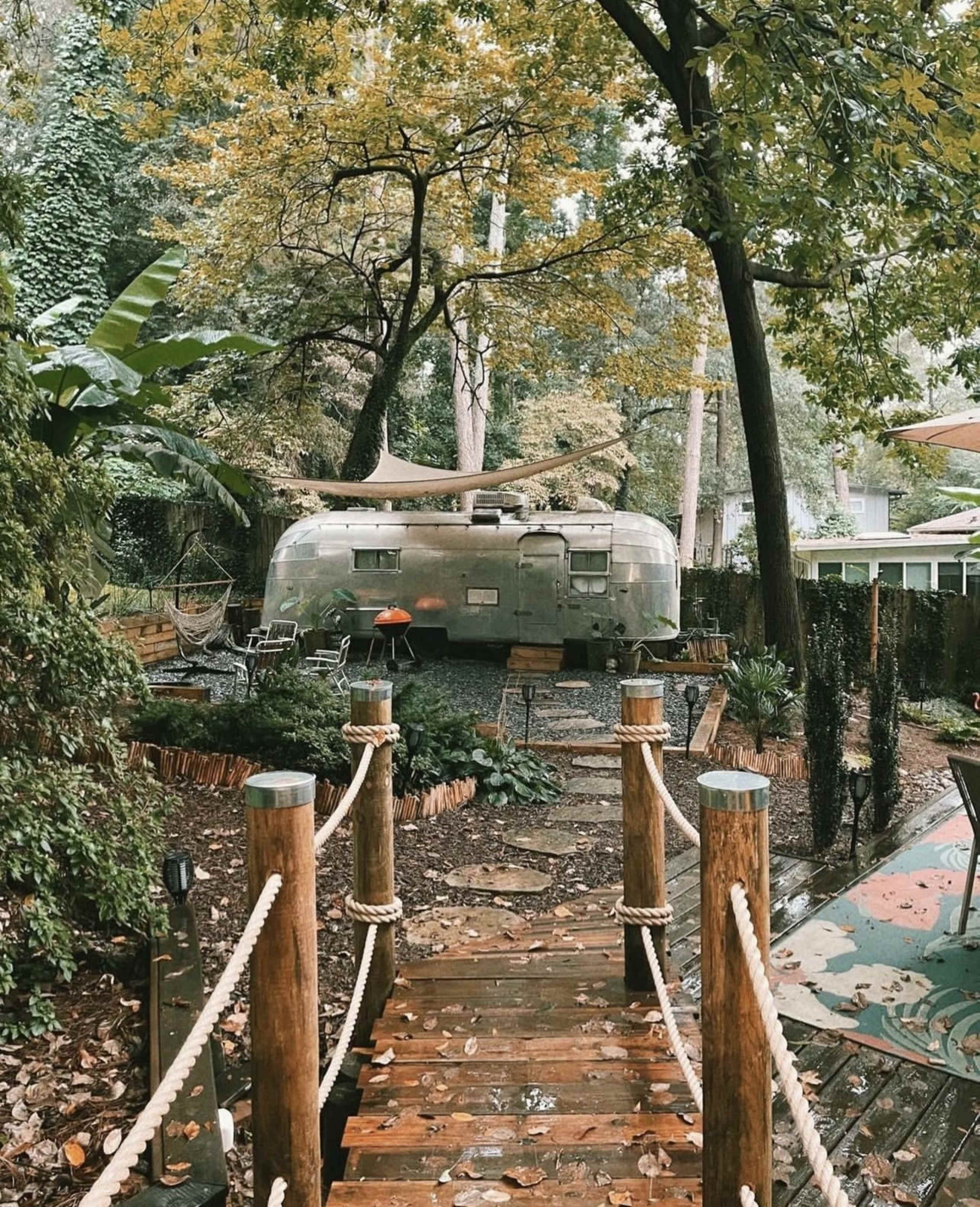A wooden pathway with rope railings leads to a vintage Airstream trailer surrounded by lush greenery and outdoor seating areas.