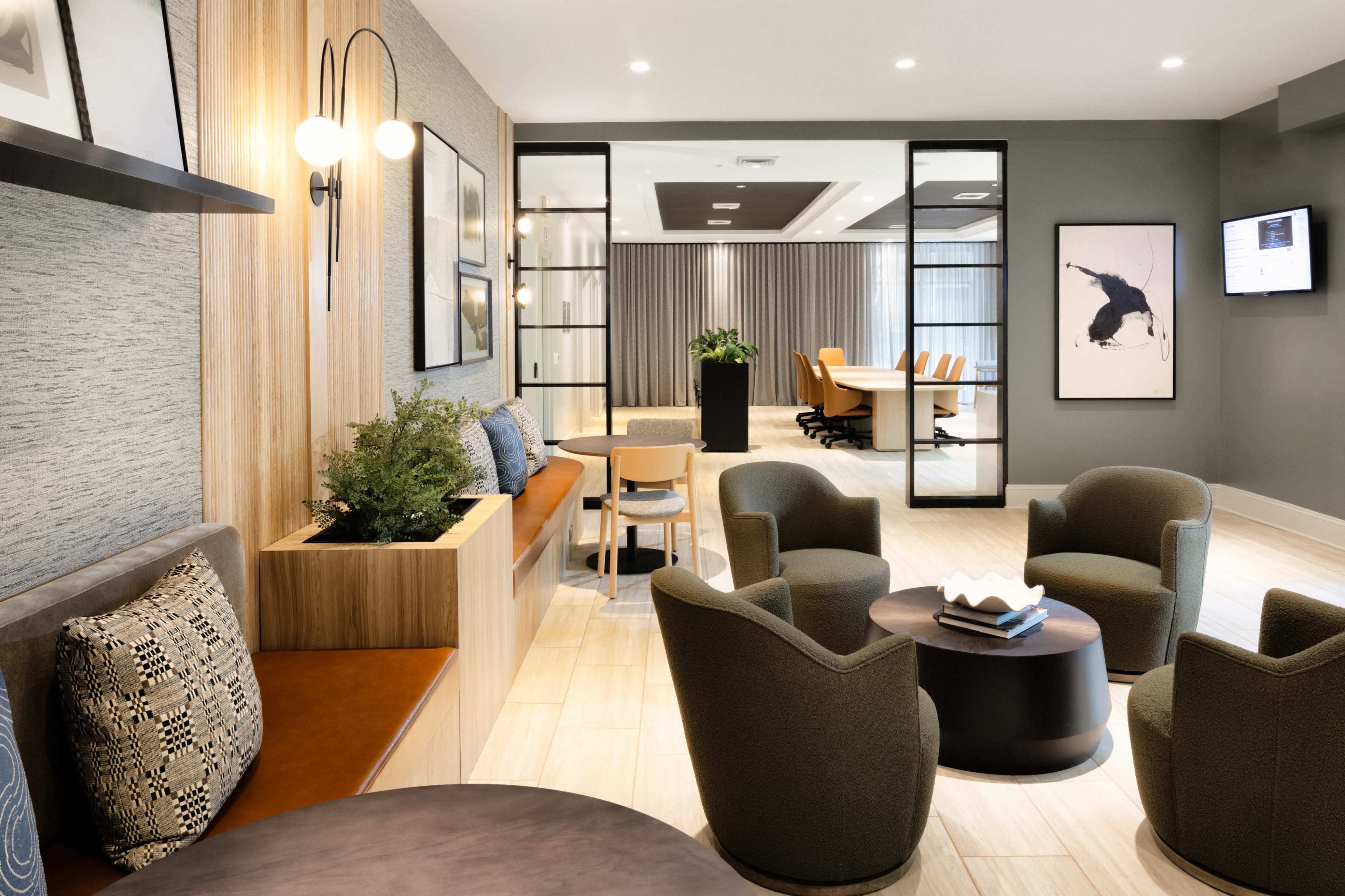 The image shows a modern lounge area with comfortable seating, a small coffee table, and a conference room visible in the background.