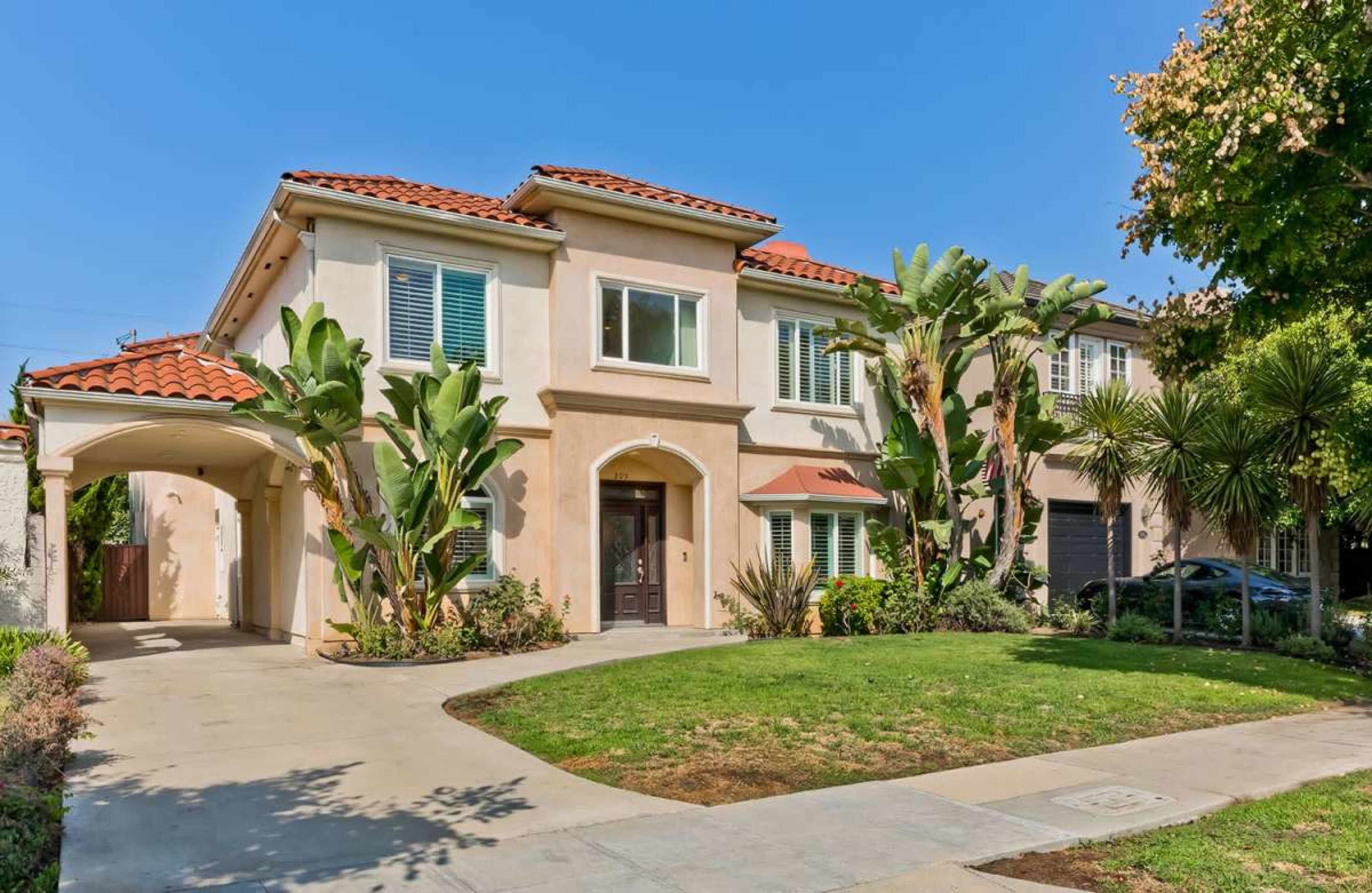 The image shows a two-story house with a tile roof, surrounded by palm trees and neatly landscaped shrubs, along a concrete walkway.