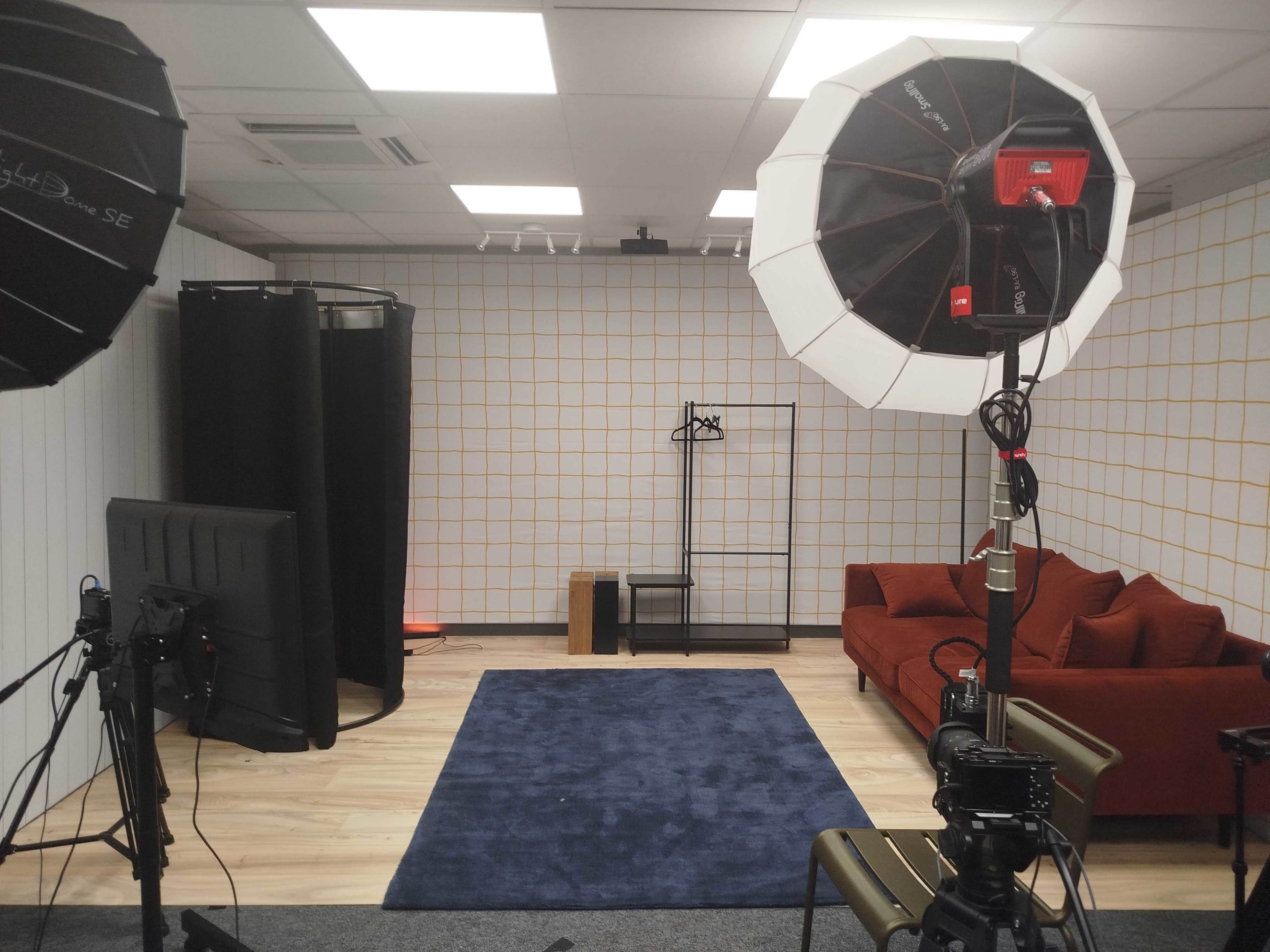The image shows a studio space with a blue rug, a brown couch, lighting equipment, and a backdrop setup featuring a grid pattern on the wall.