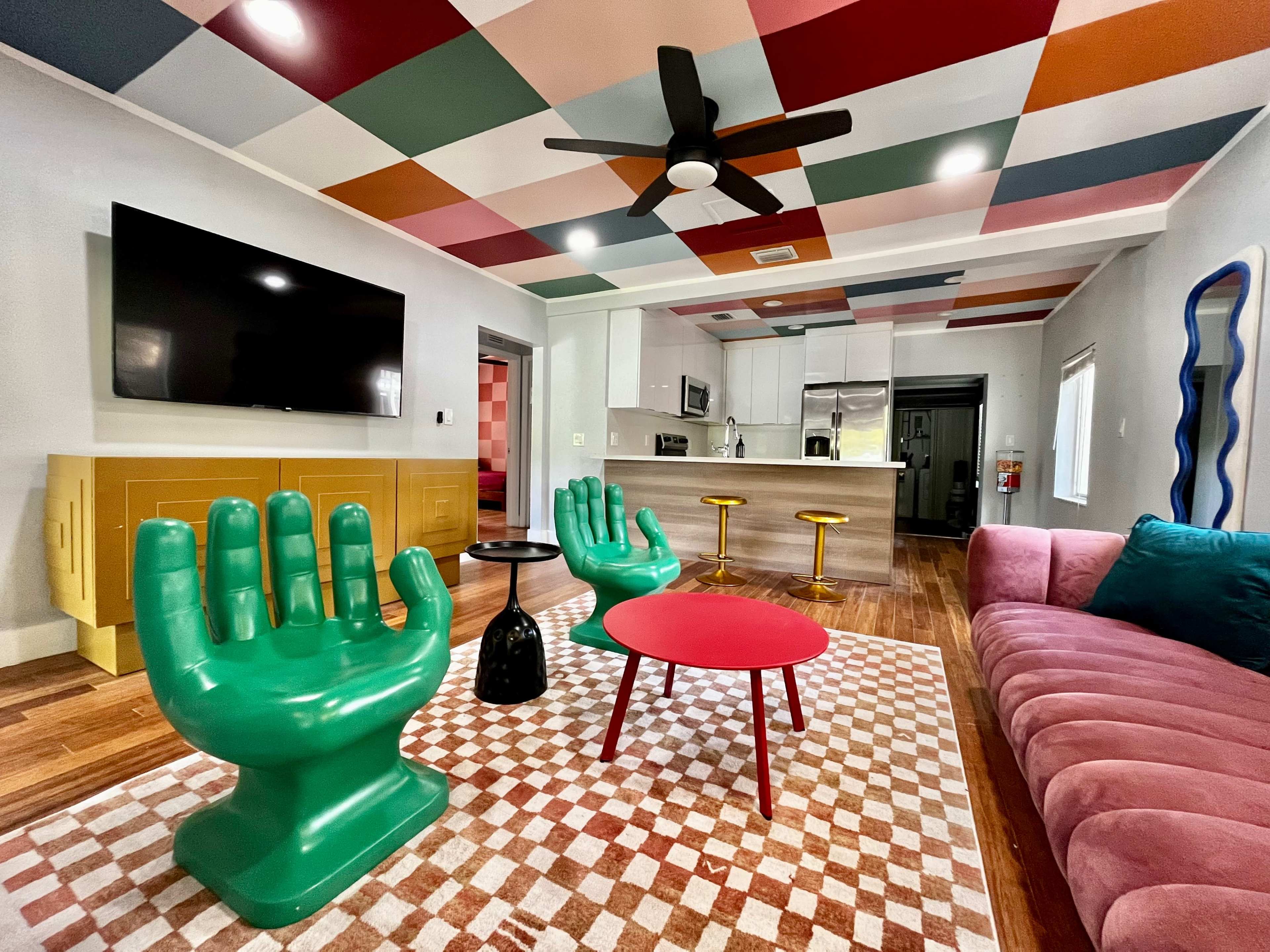 A vibrant living room featuring bright green hand-shaped chairs, a pink round coffee table, and a sofa, with a colorful patterned ceiling and an open kitchen in the background.