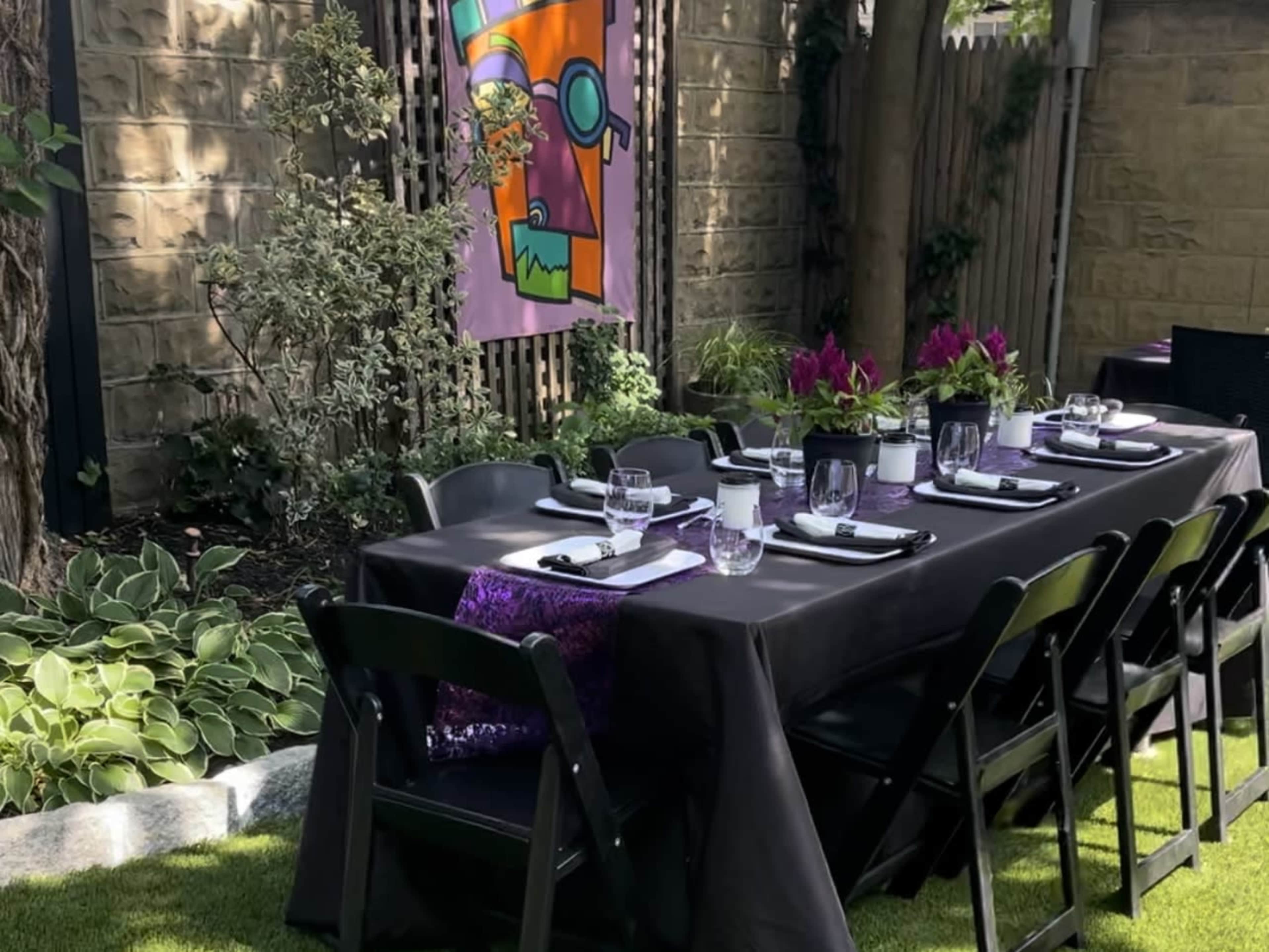 A long black table is set for dining in a garden, surrounded by greenery and a colorful banner hanging nearby.