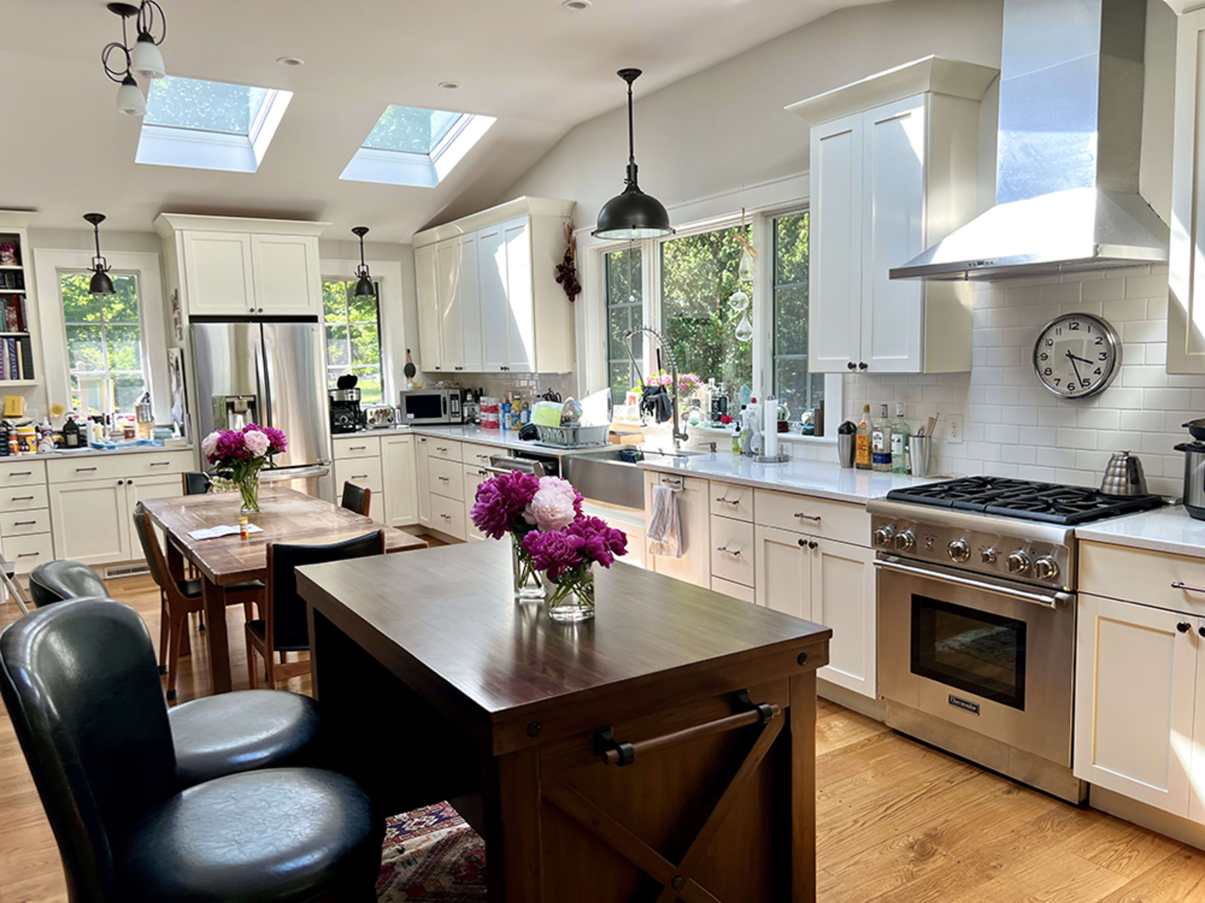A spacious kitchen features white cabinetry, a large island with a wooden table, and skylights that illuminate the room.
