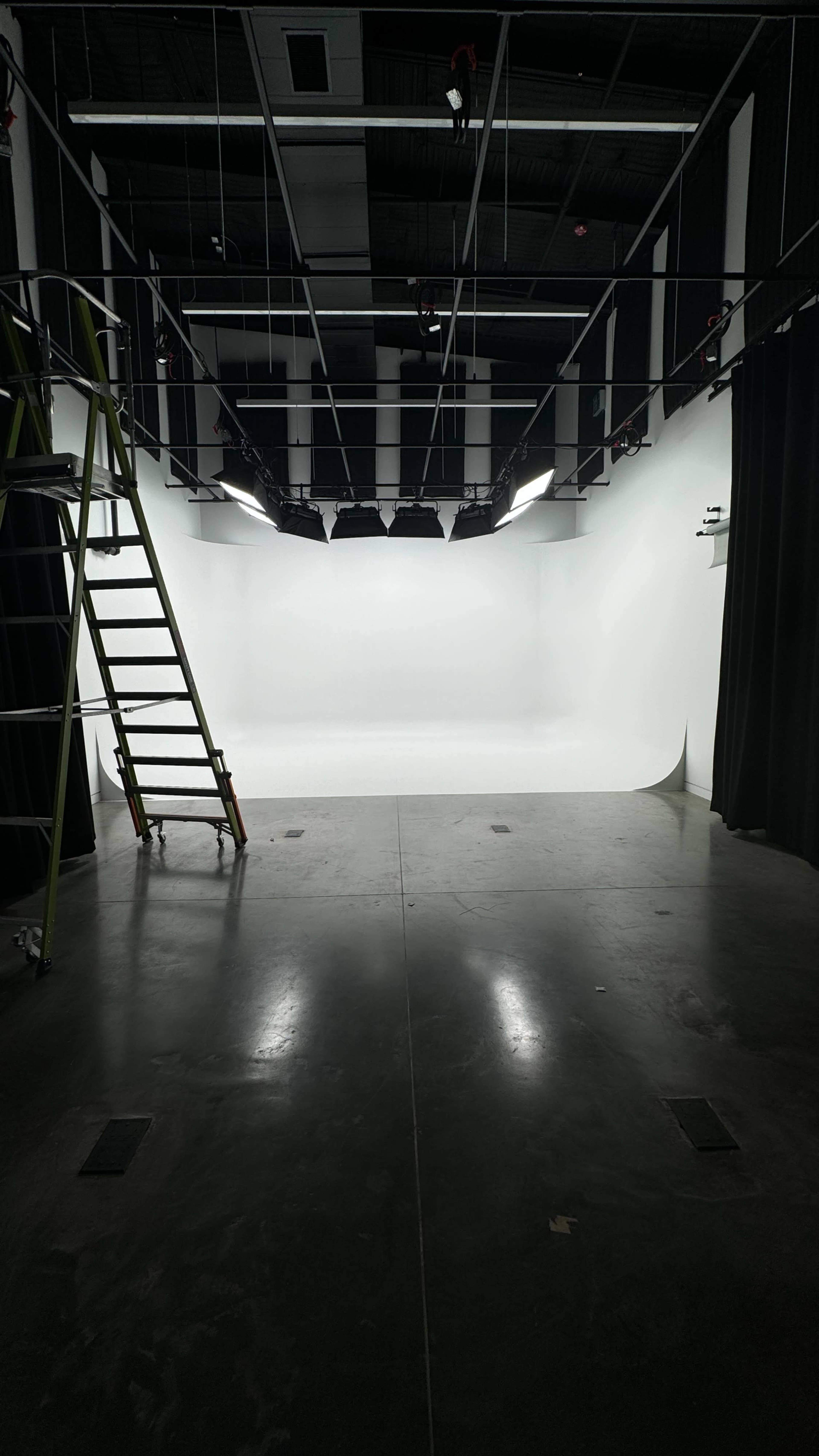 The image shows a photography studio with a ladder on the left side and a seamless white backdrop in the center, illuminated by overhead lights.