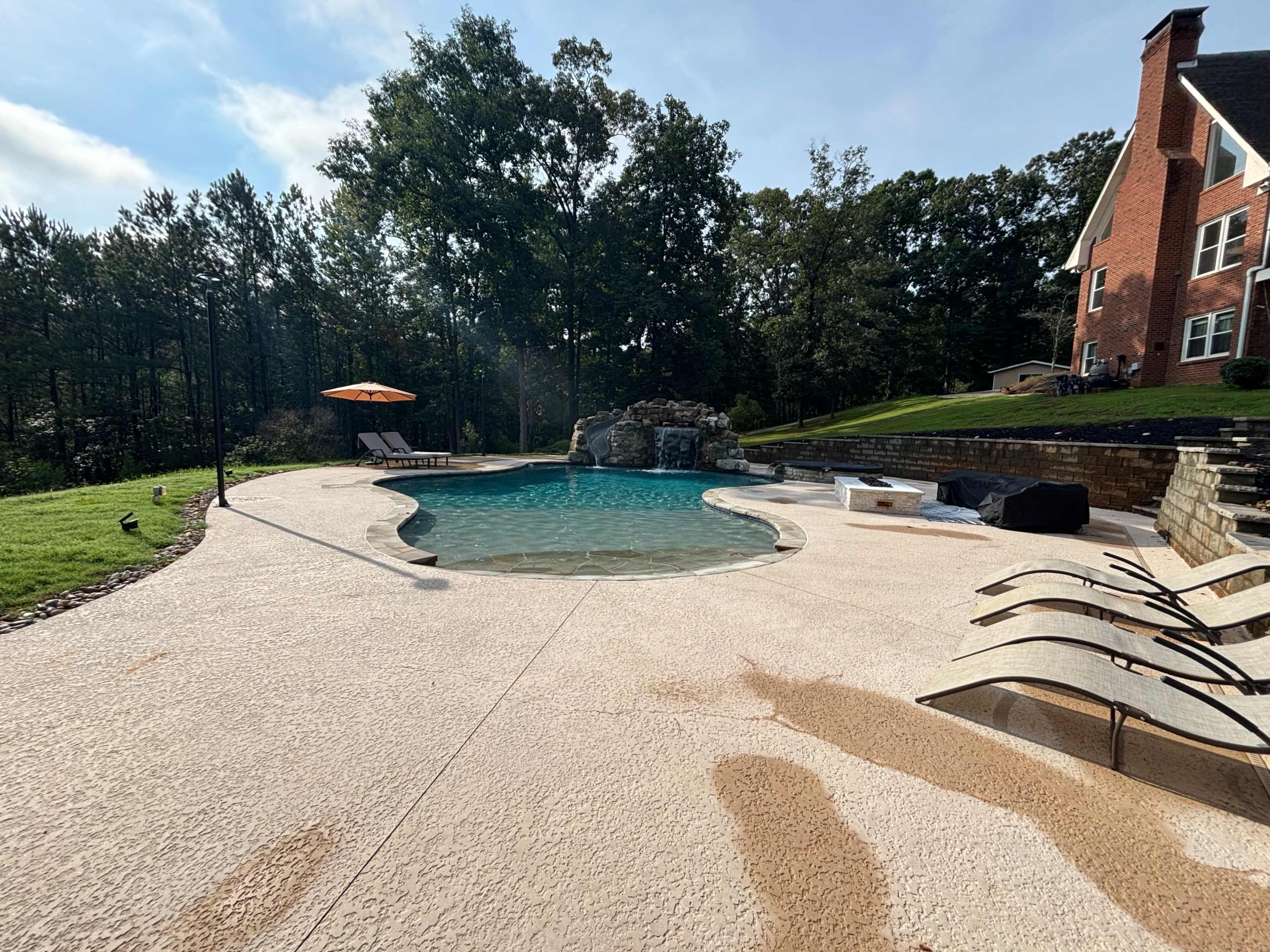 The image shows a pool area featuring a curved swimming pool, lounge chairs, a rock waterfall, and a nearby house set against a backdrop of trees.