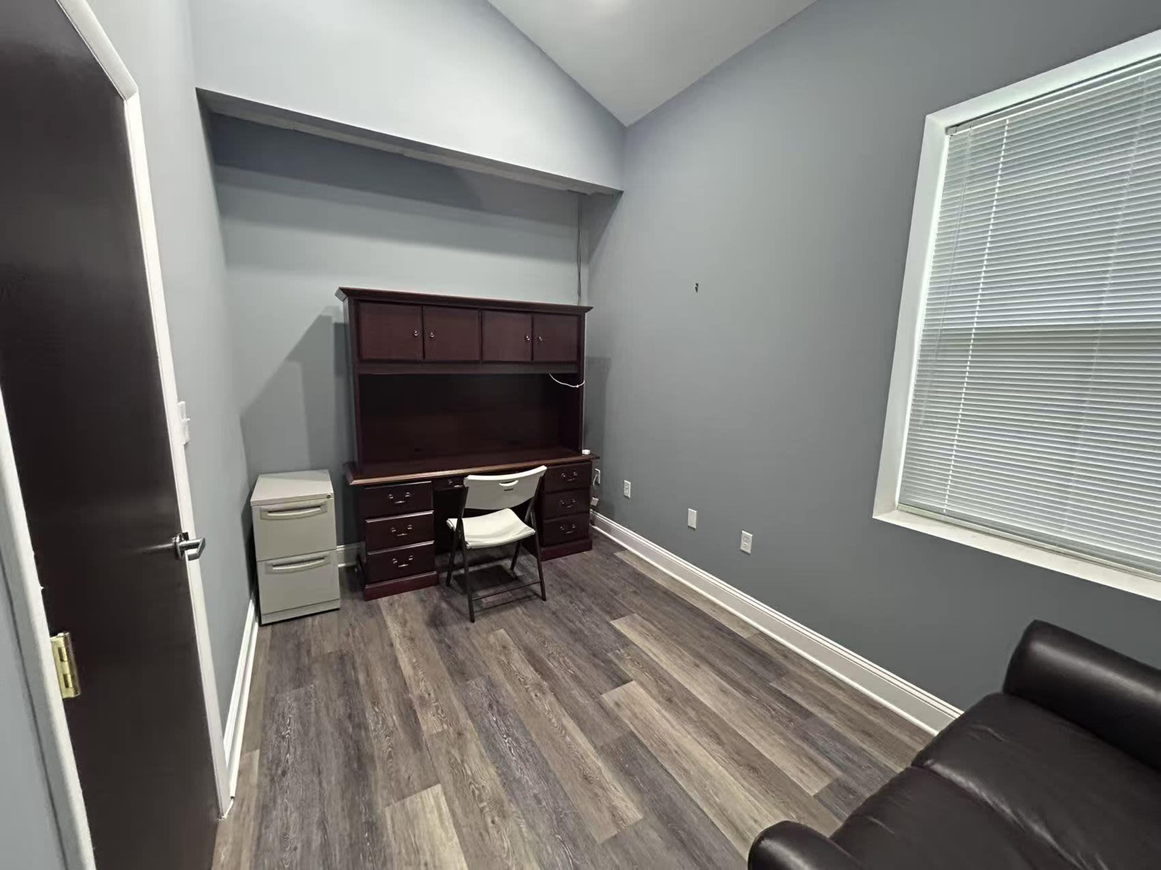 The image shows a small office space featuring a wooden desk with a chair, a filing cabinet, and a couch against the wall, with gray walls and a window.