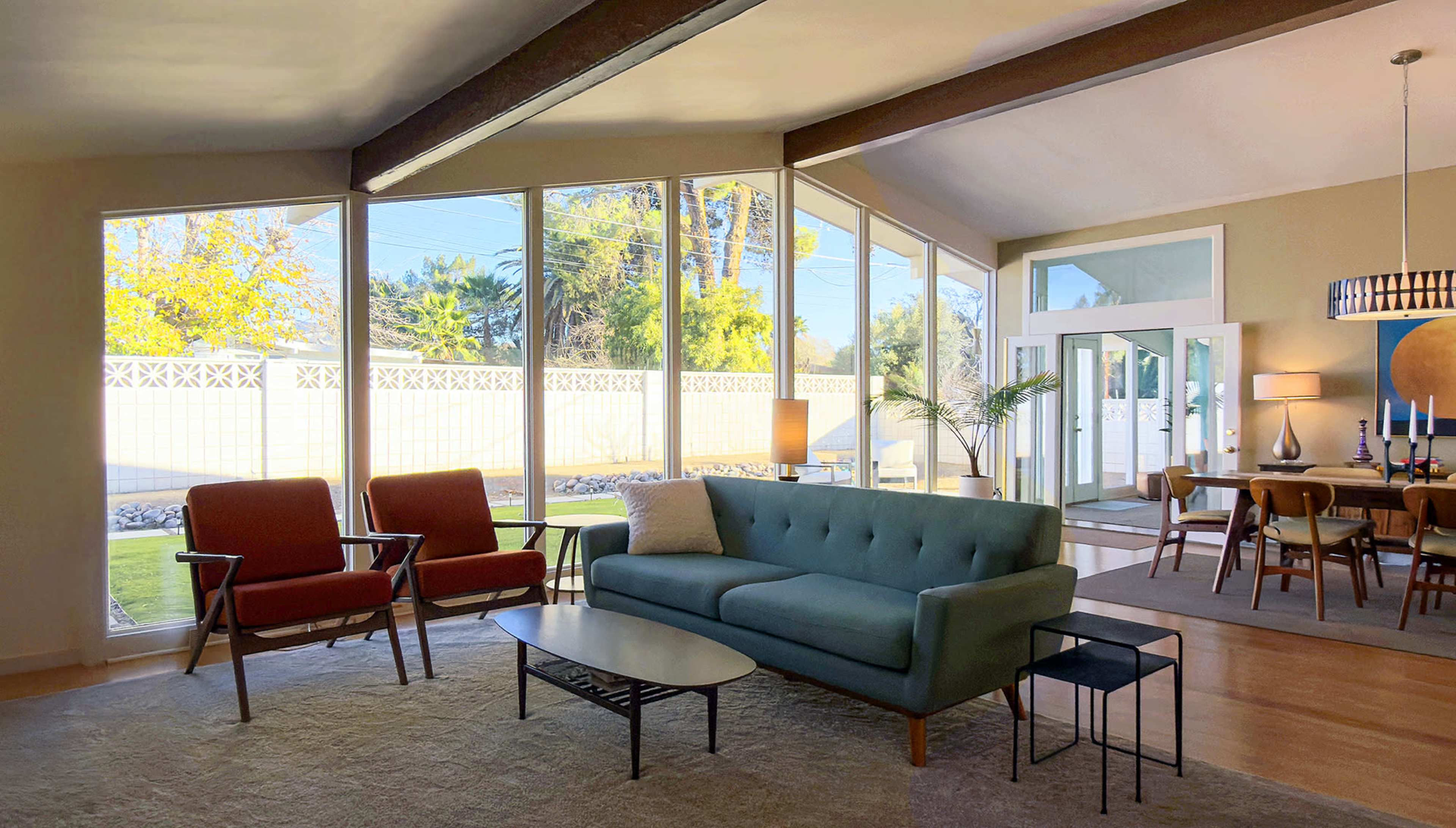 The image shows a modern living room with a blue sofa, two red chairs, and large windows that provide a view of a garden outside.