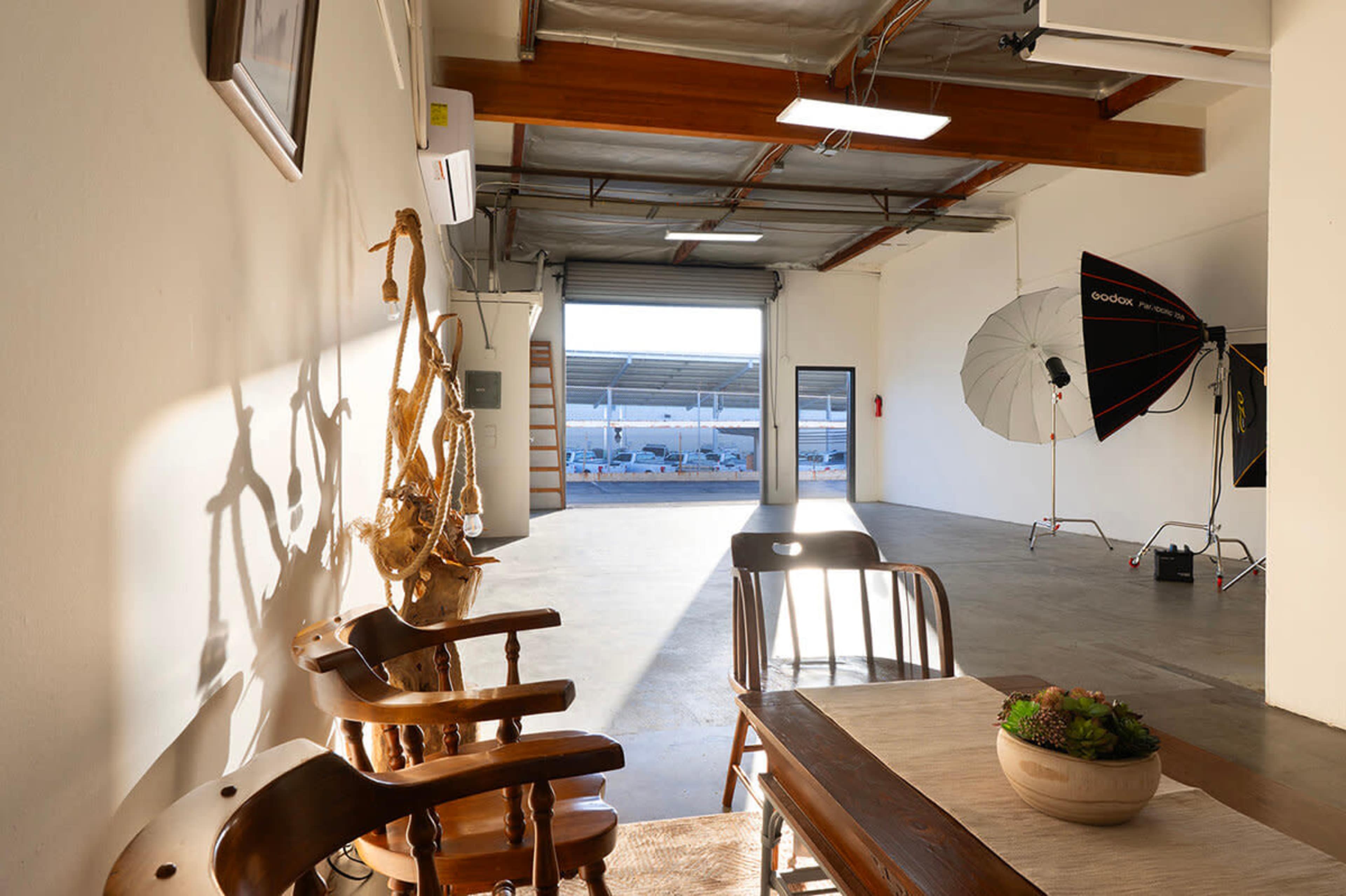 The image shows a spacious, well-lit studio or garage featuring a wooden dining table, chairs, and photography equipment near an open bay door.