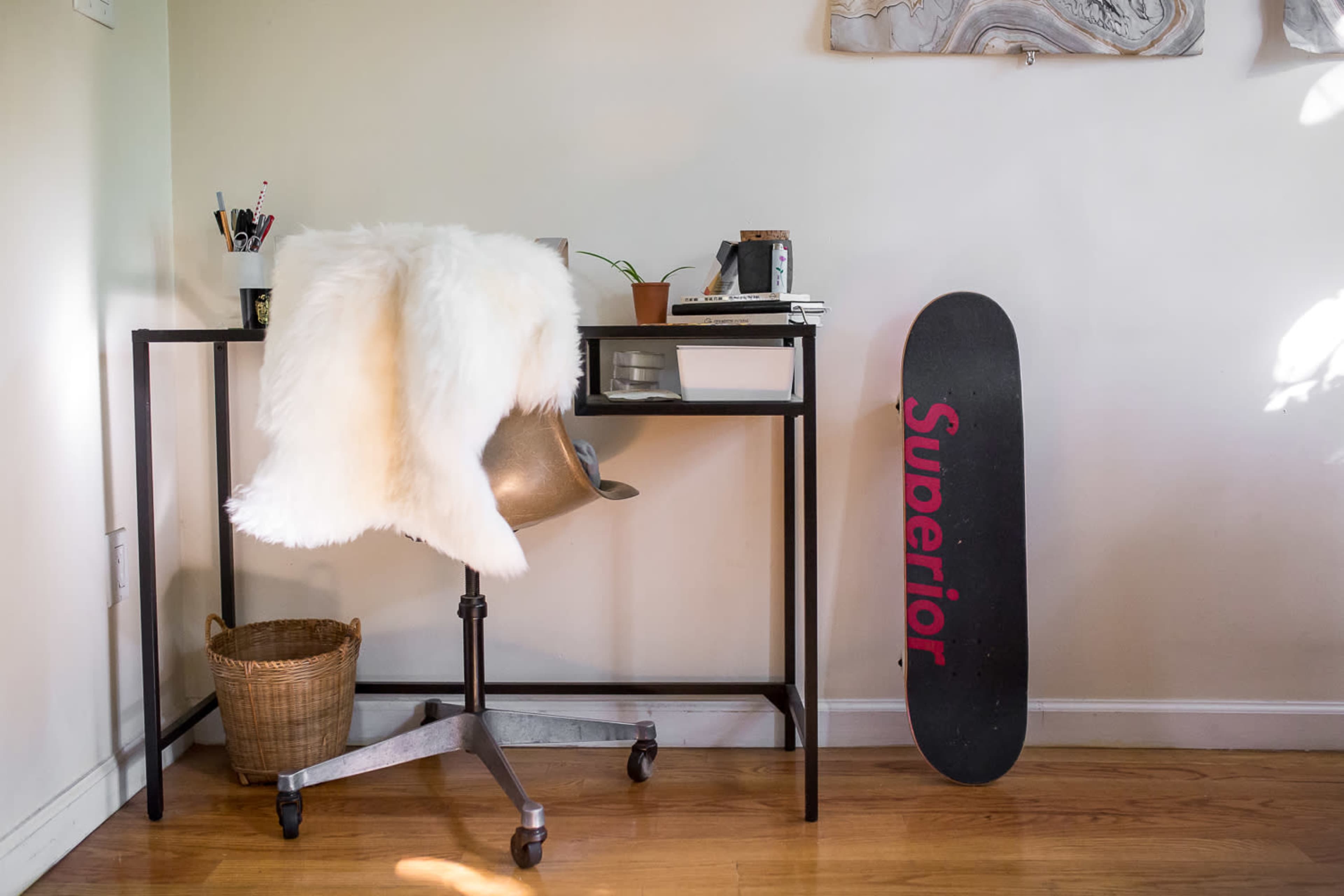 A modern workspace features a black metal desk with an ergonomic chair, a fur throw, a potted plant, and a skateboard propped against the wall.