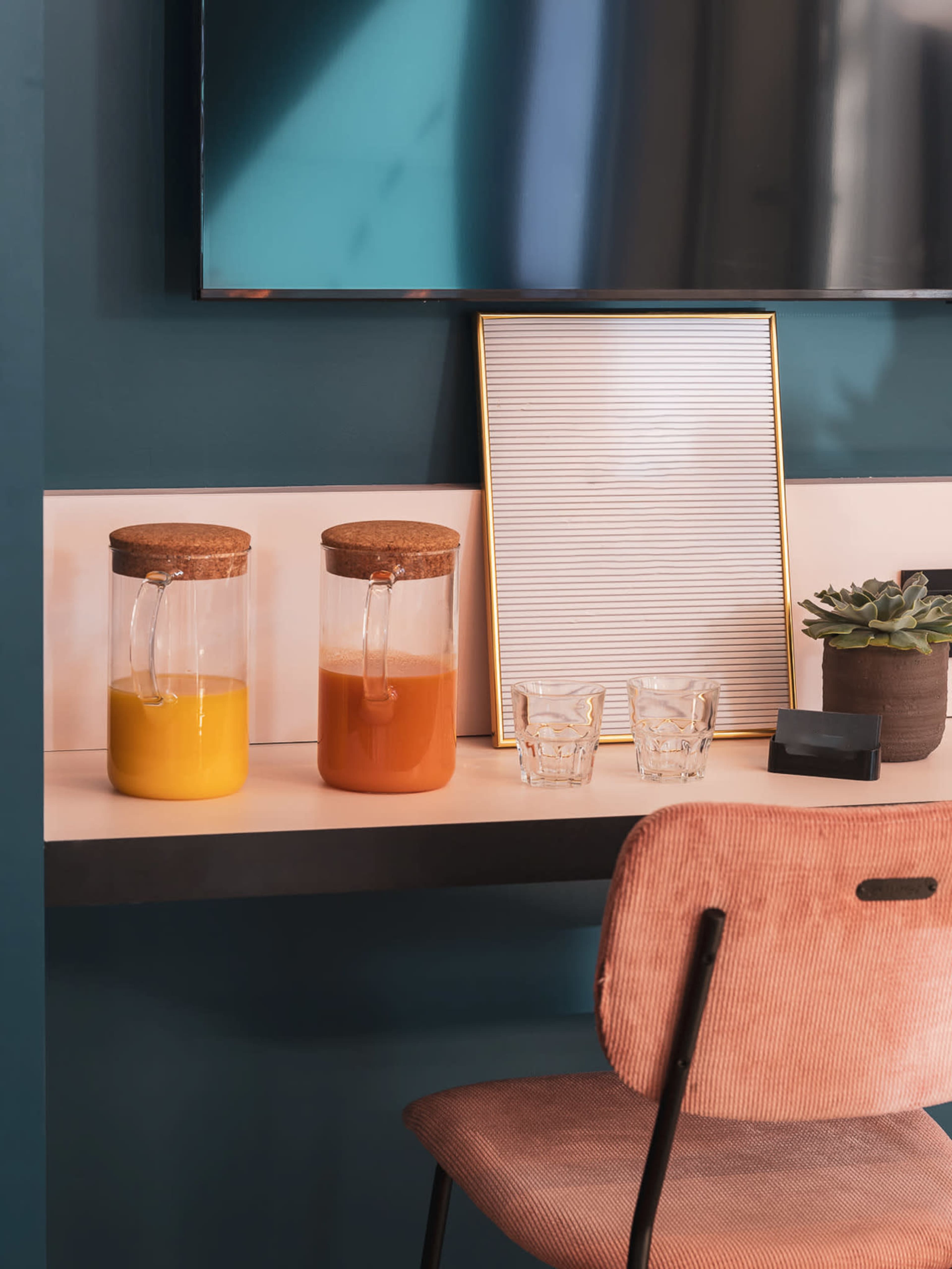 The image shows a countertop with two glass pitchers of yellow and orange liquids, a small succulent plant, two clear glasses, and a framed decorative board on a teal wall.
