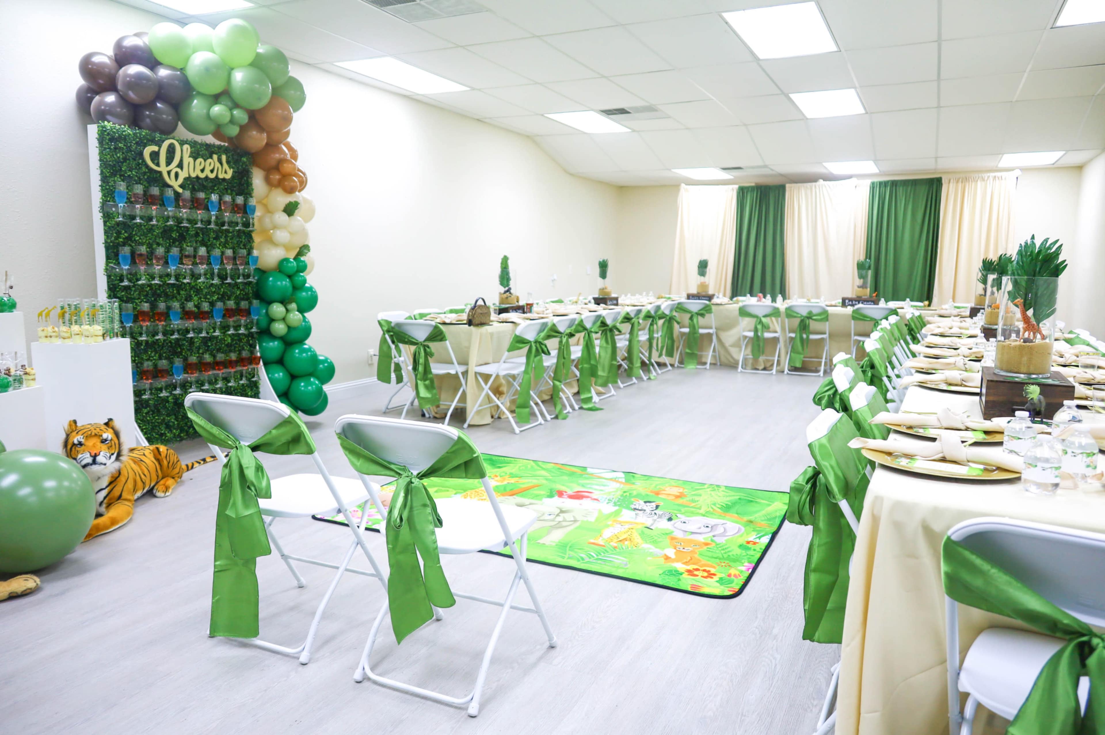 A decorated event space with tables set for a gathering, featuring green and brown balloons, floral arrangements, and a themed jungle rug.