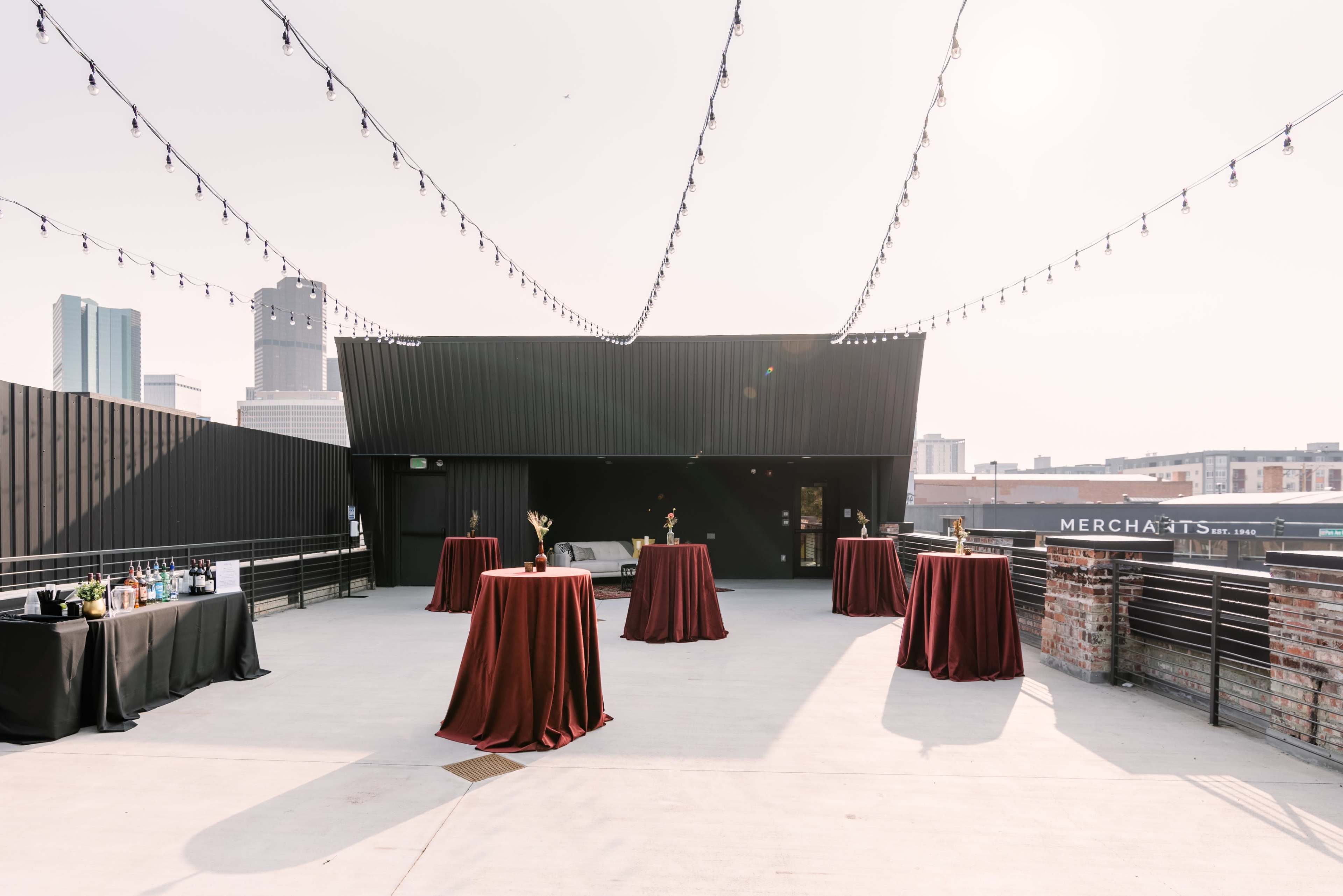 The scene depicts a rooftop event space with several round tables covered in maroon tablecloths, string lights overhead, and a city skyline in the background.