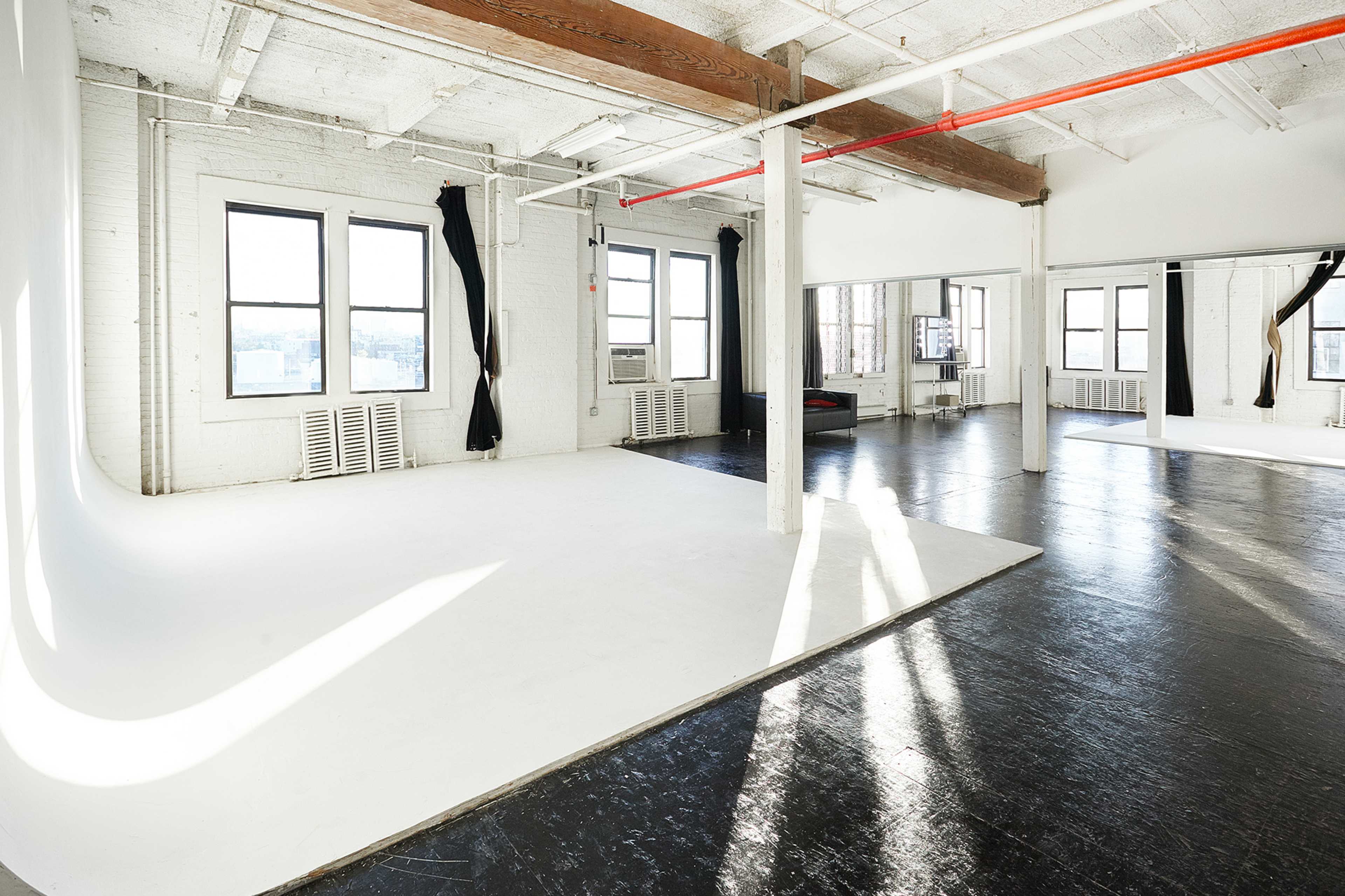 A spacious, industrial-style room with white walls, large windows, and a black polished floor, featuring a seamless backdrop area on one side.