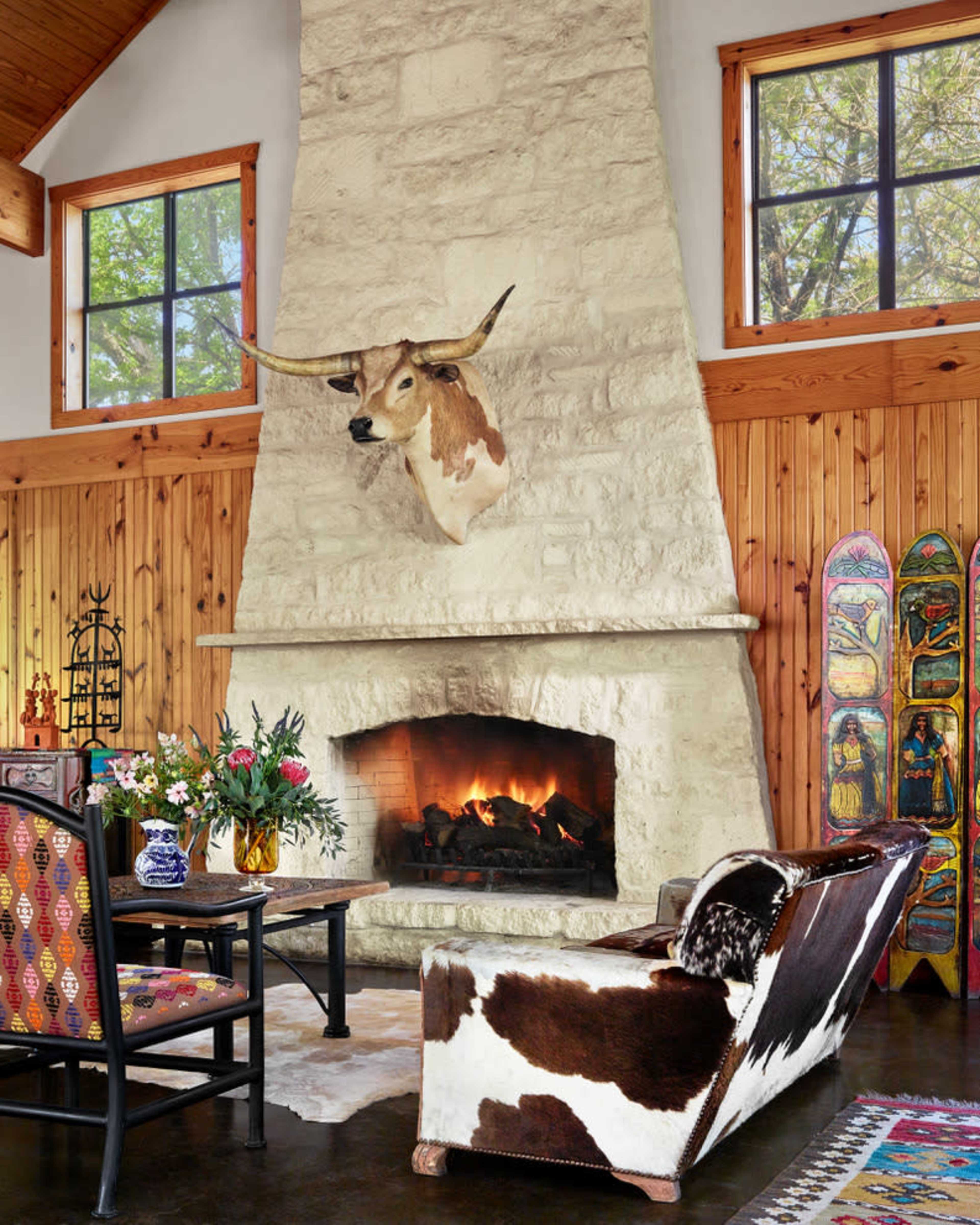 A cozy living room features a stone fireplace with a mounted longhorn head above it, surrounded by wooden walls, a patterned rug, and cowboy-themed decor.
