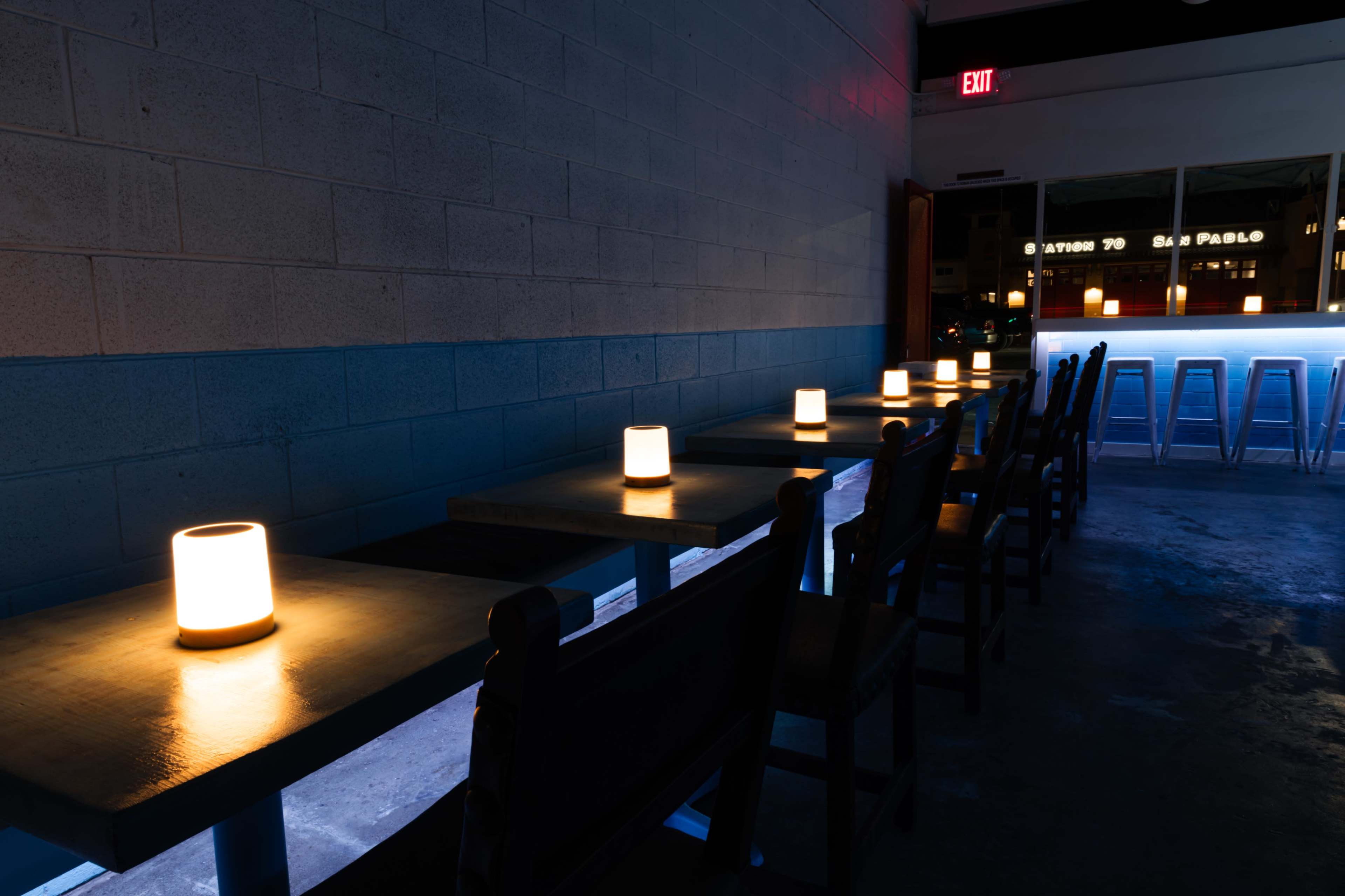 The image shows an interior space with several tables illuminated by glowing cylindrical lamps, creating a dimly lit atmosphere.