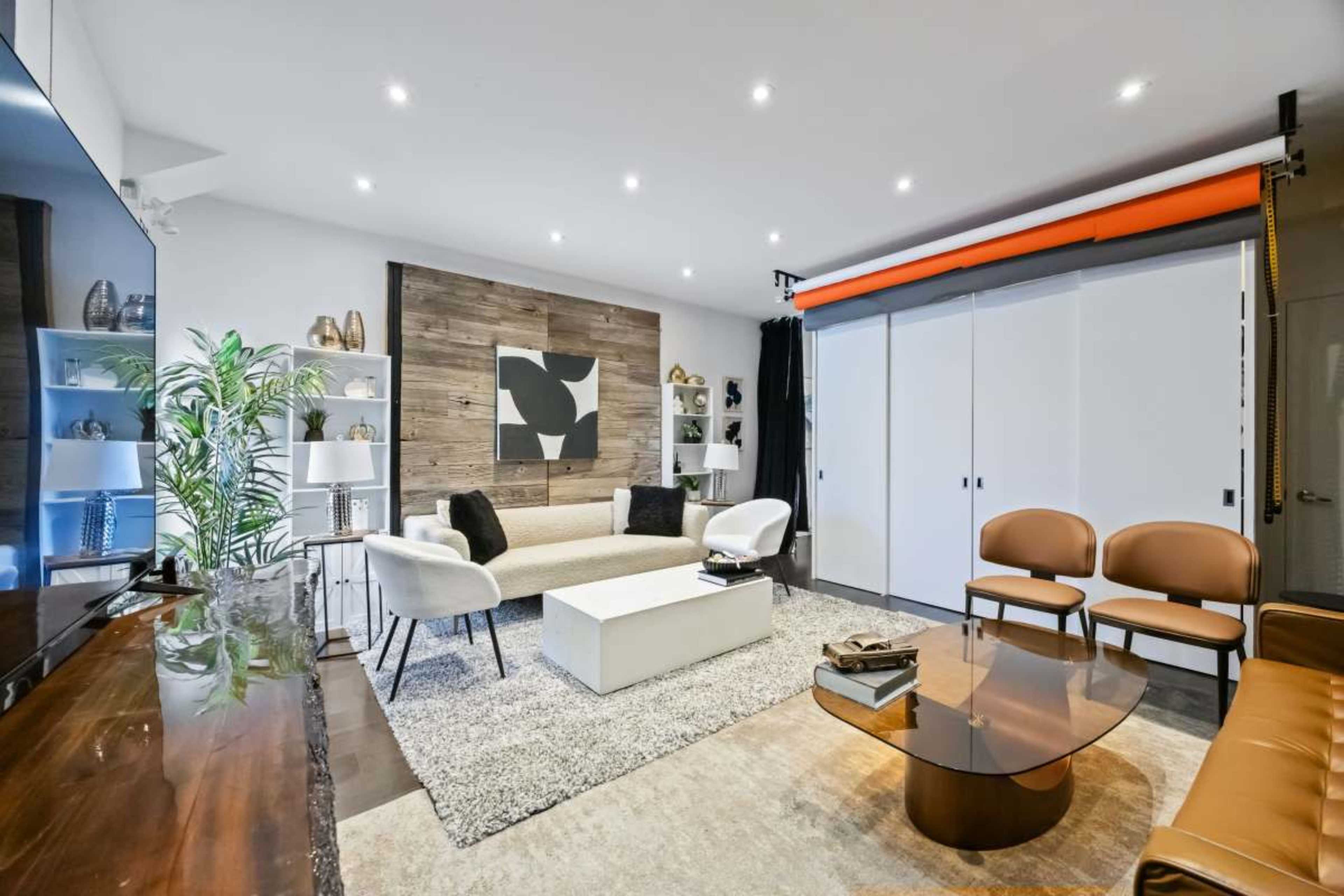 The image shows a modern living room with a white sofa, two brown chairs, a coffee table, and a wall featuring decorative elements and a large piece of art.