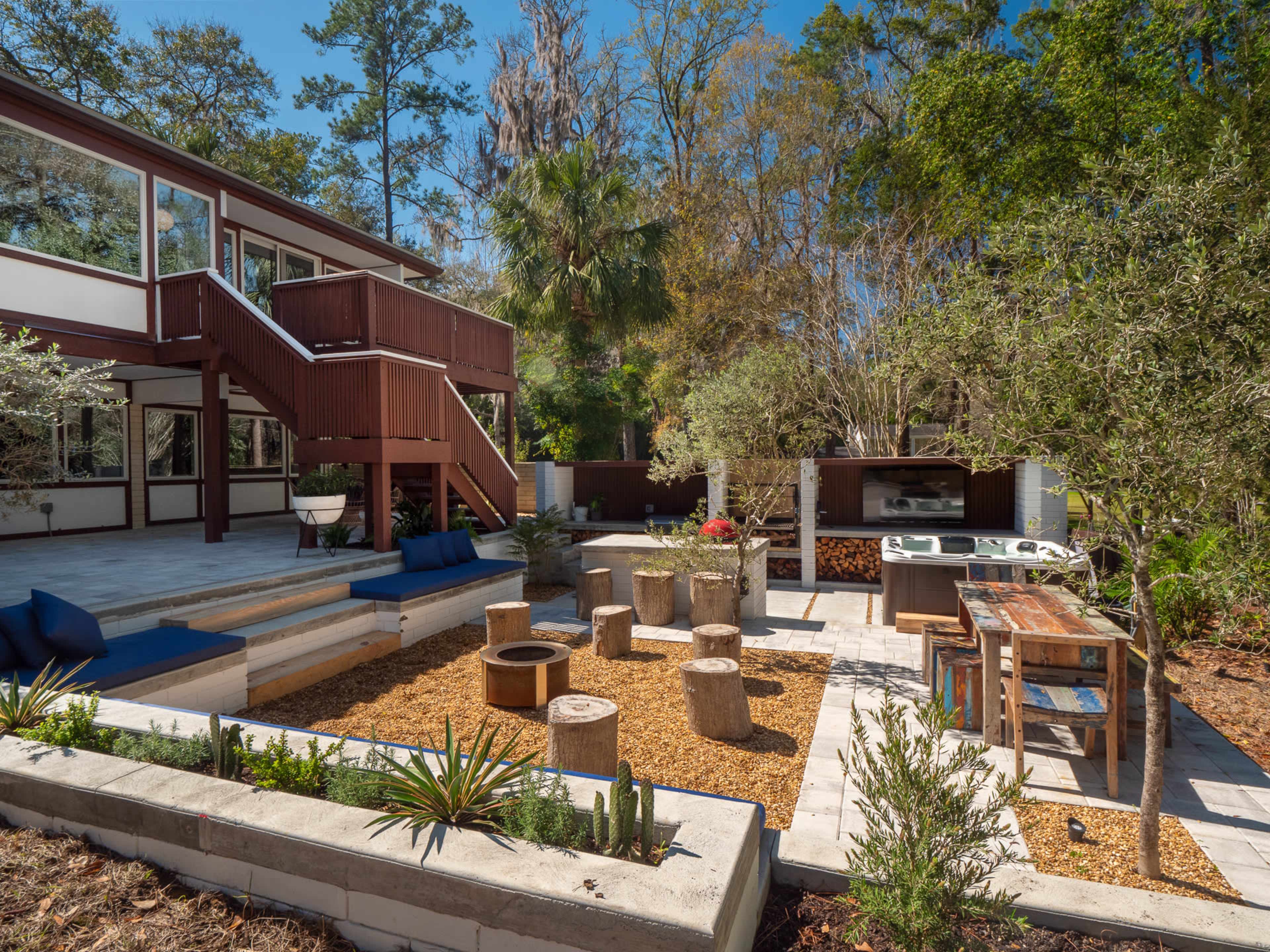 The image shows a landscaped backyard area featuring a stone patio, wooden seating around a fire pit, an outdoor kitchen, and a multi-level home in the background.
