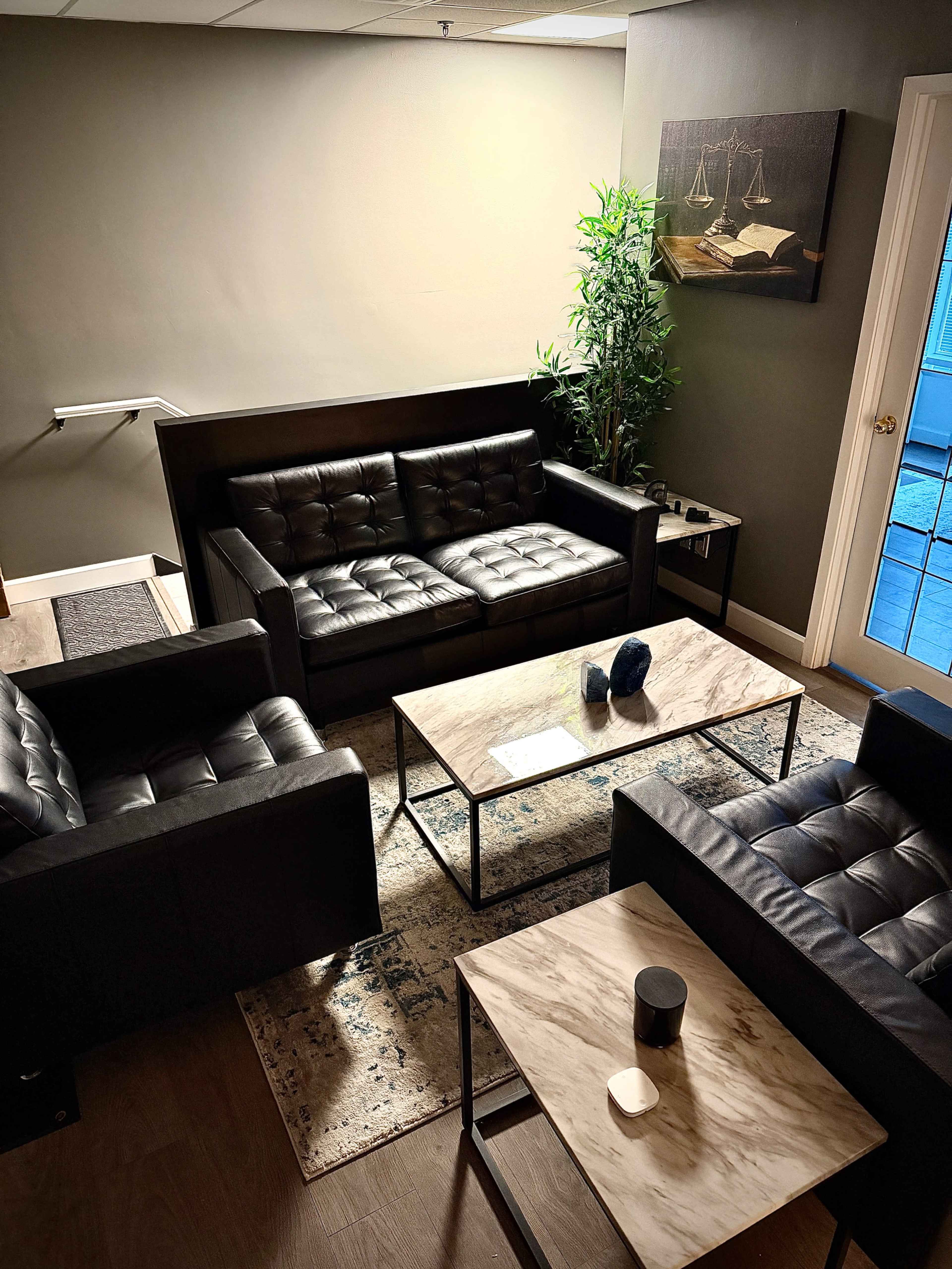 The image shows a modern lounge area furnished with black leather sofas and a marble top coffee table, complemented by a potted plant and a painting on the wall.