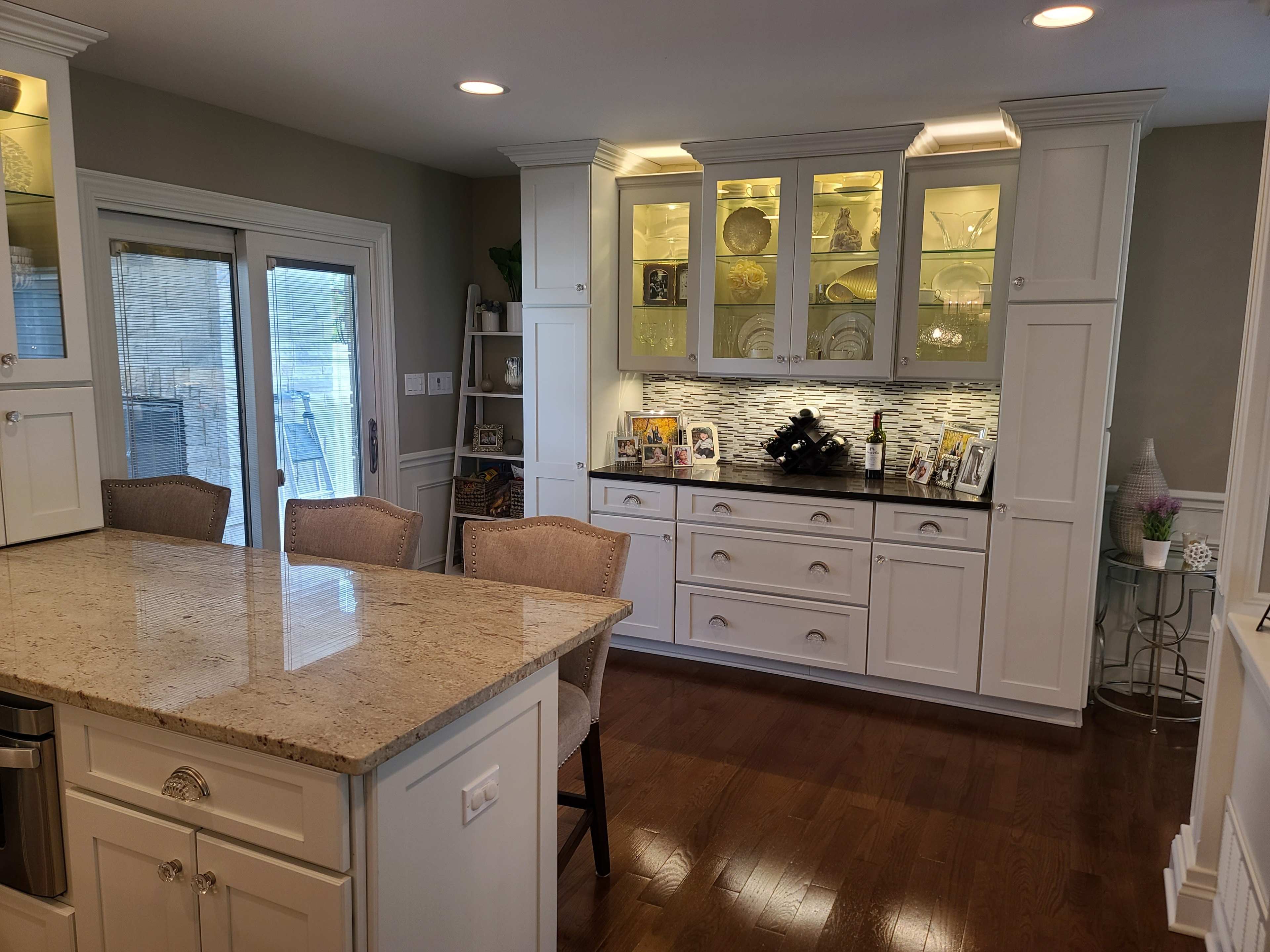 A modern kitchen with white cabinetry, a granite countertop island, and glass-front cabinets displaying decorative items.