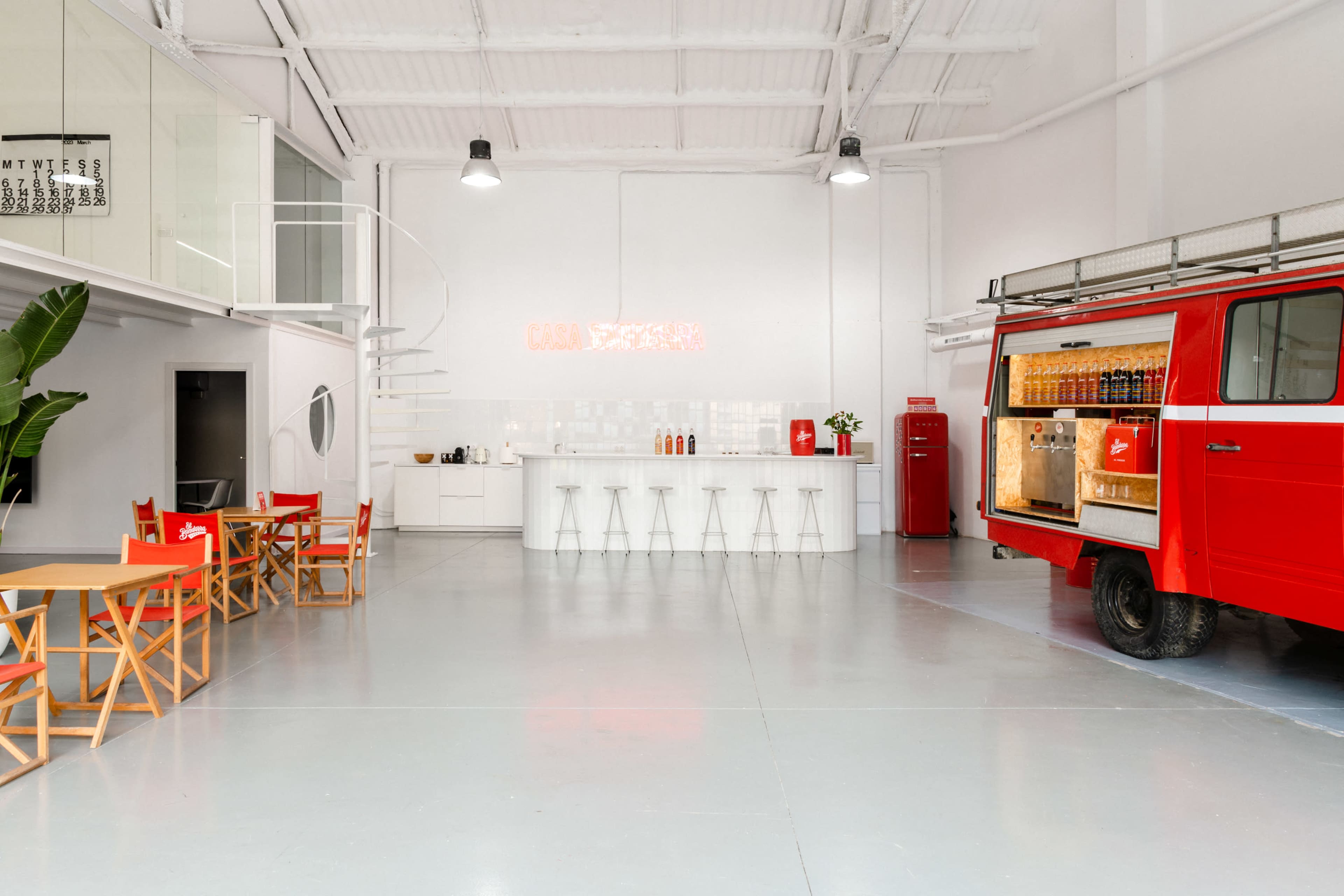A spacious interior features a modern kitchen area with a bar counter, a red vintage van, and wooden tables arranged for seating.