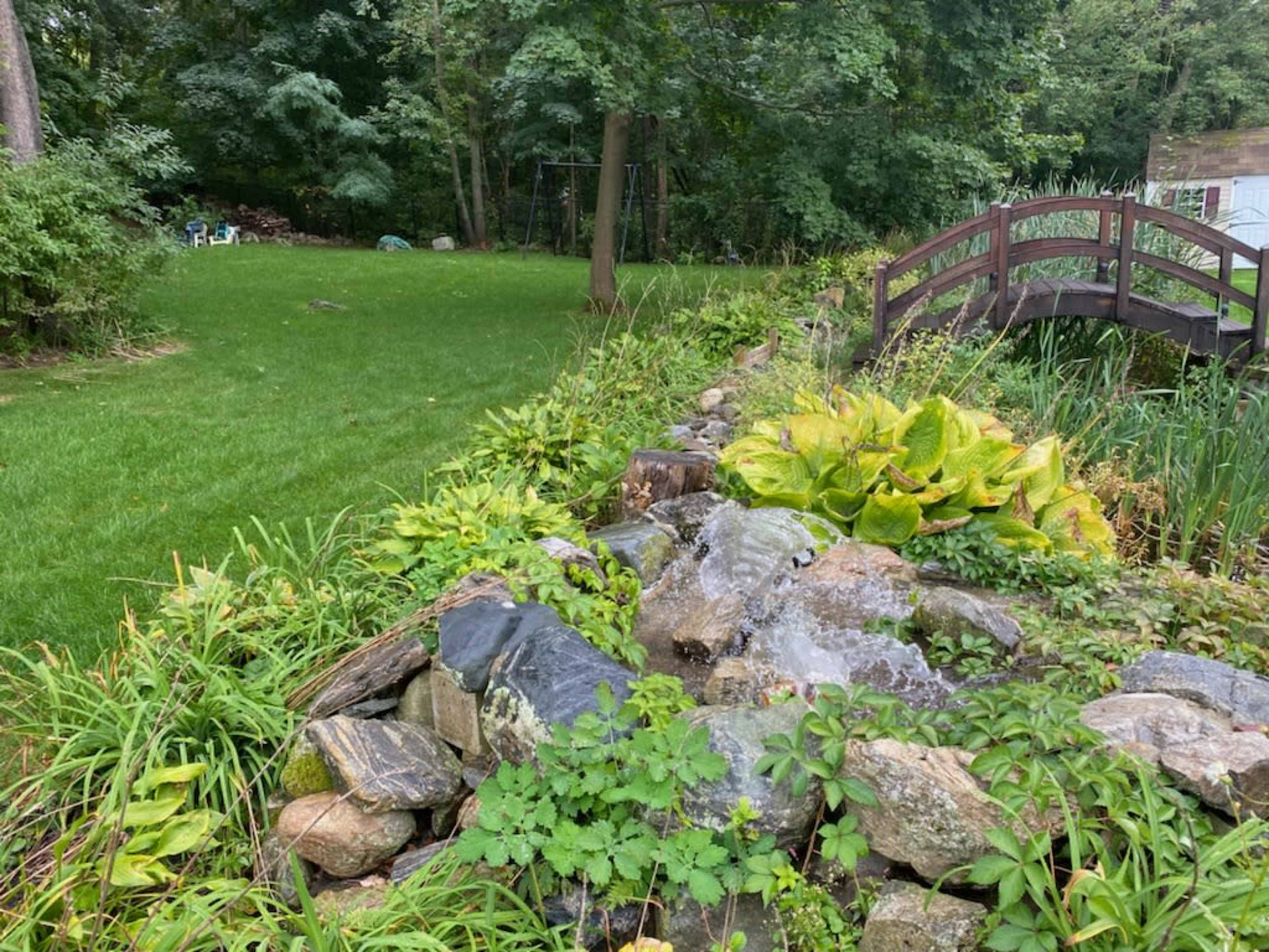 A small waterfall cascades over rocks beside a lush green lawn and wooden bridge in a landscaped garden.