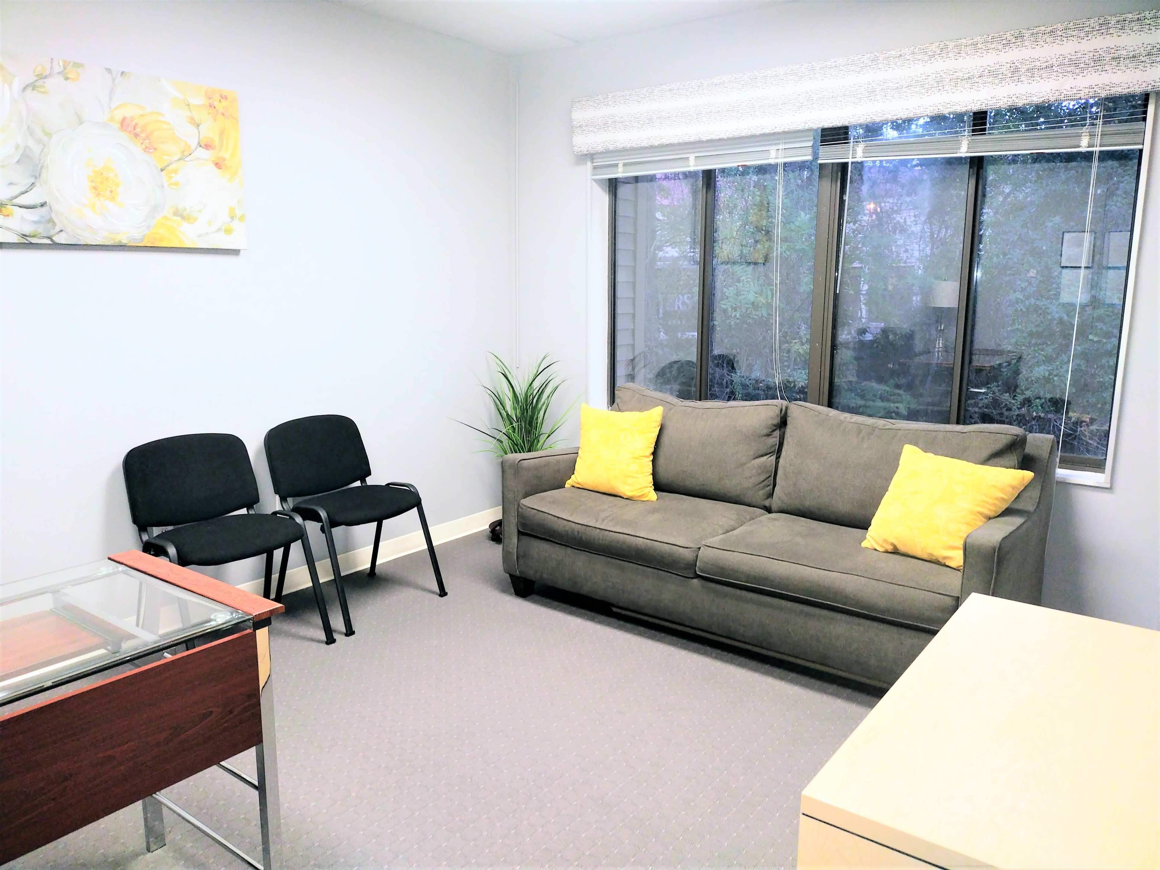 A small office with a sofa, two chairs, a glass table, and a window overlooking greenery.